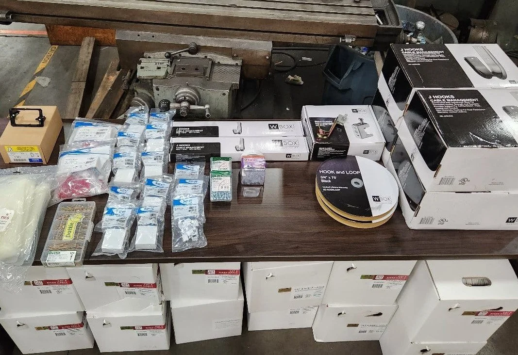 Assorted hardware and tools, including boxes of cable hooks, wire, and accessories, arranged on a workshop table with storage boxes underneath.