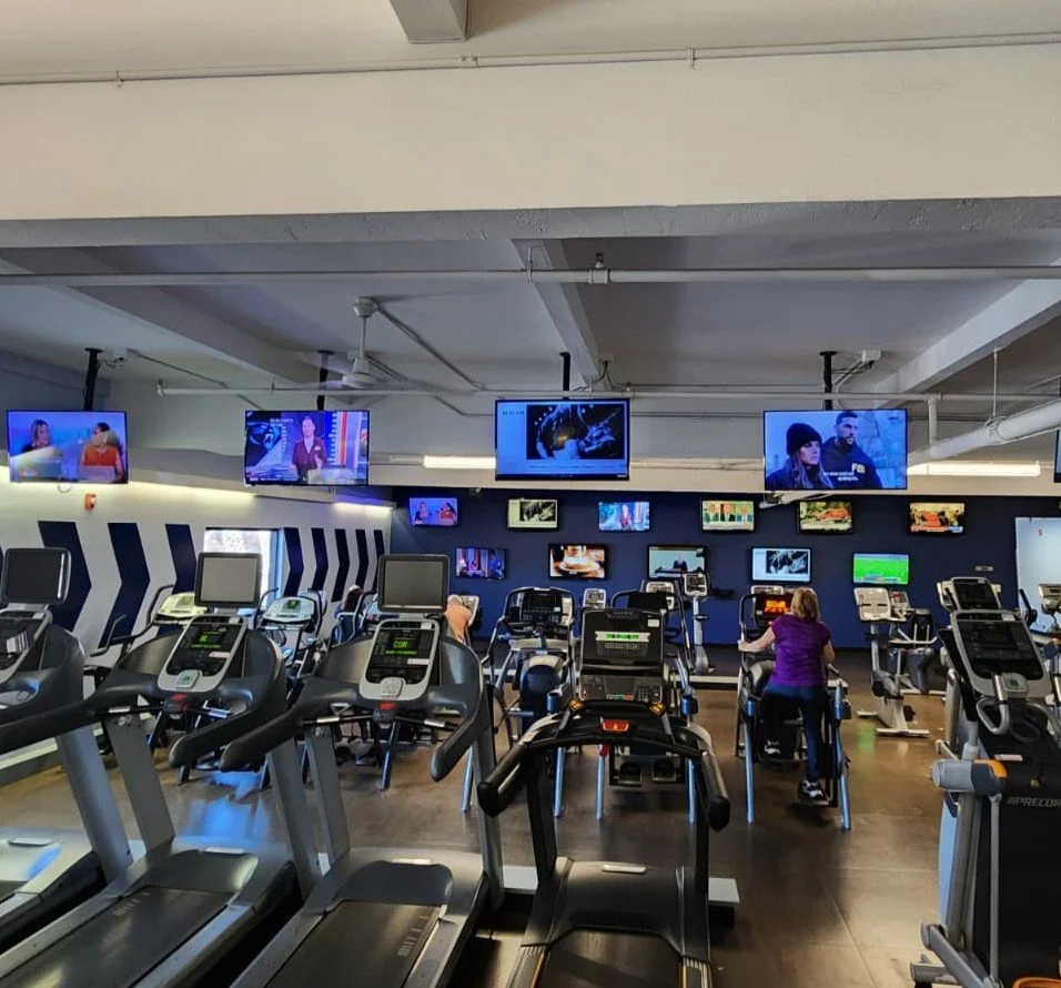 An indoor gym with multiple treadmills facing a wall of television screens, and a few people using the equipment.