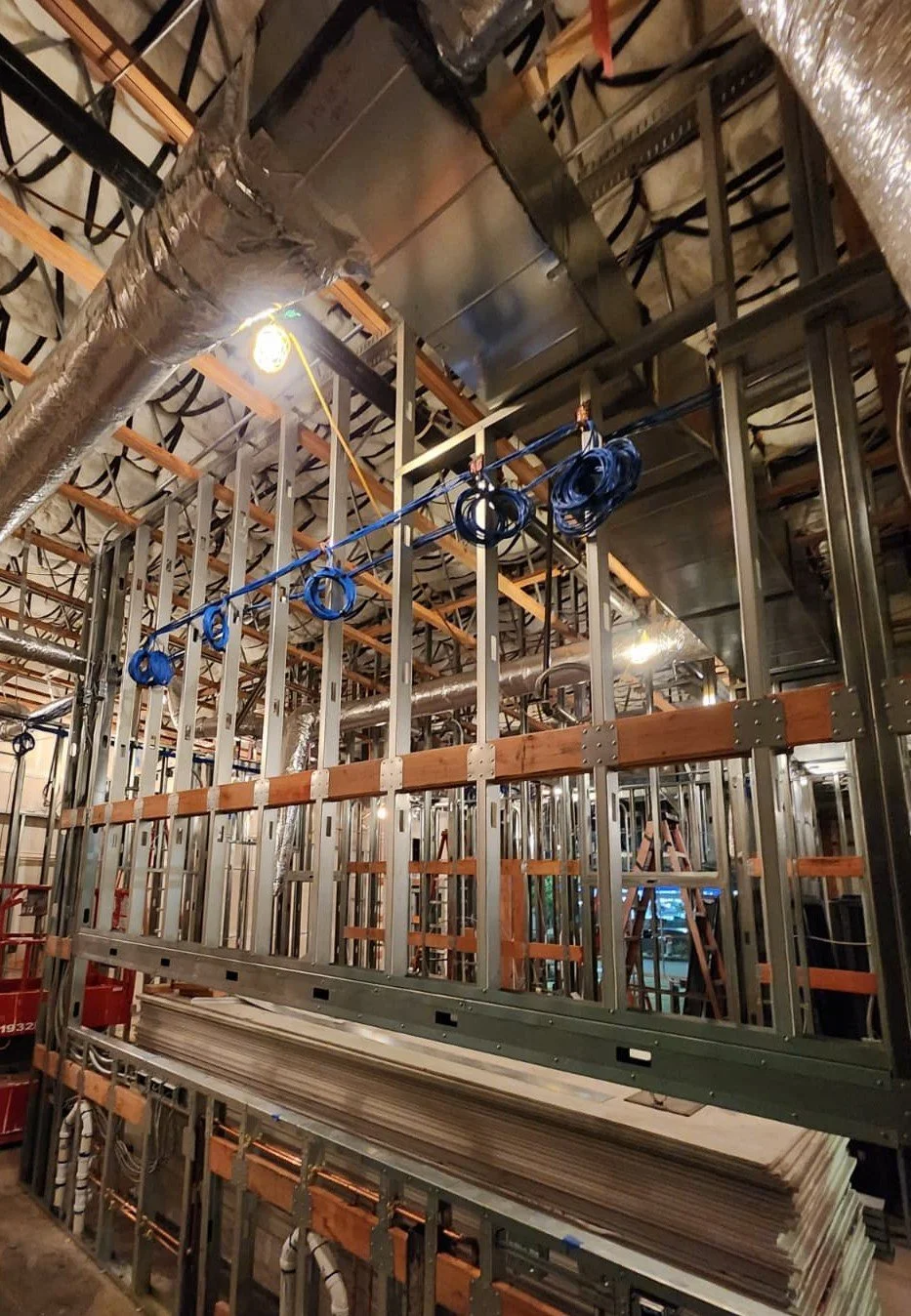 Construction site with metal framing, wooden beams, ductwork, and blue cables hanging from the ceiling.