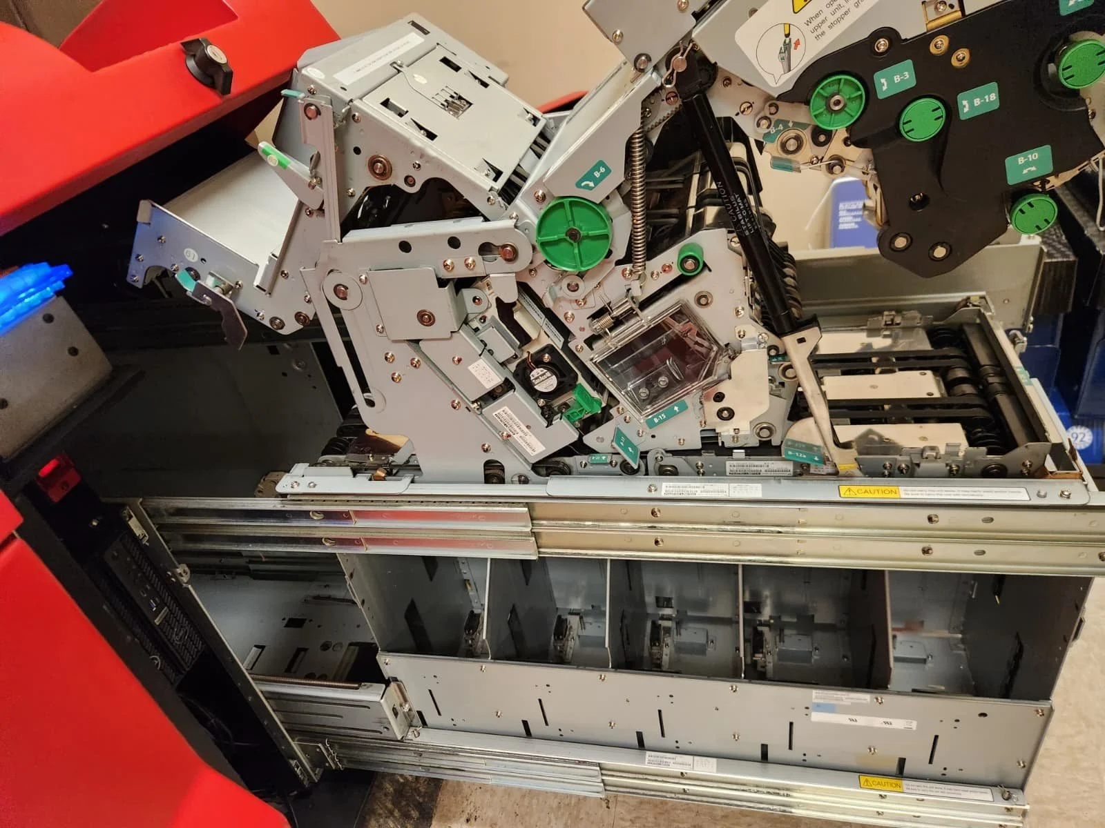 The image shows an open mechanical device, possibly a piece of industrial or manufacturing equipment, with various components, gears, and green circular labels.