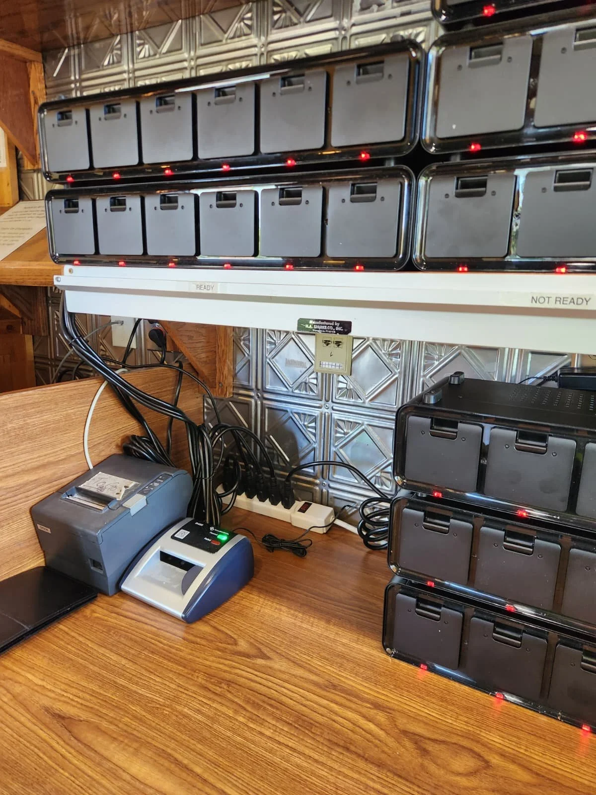 Multiple black battery packs with red LED lights are mounted on a wall rack. A desk with a receipt printer, a card reader, and connected cables is visible in the foreground.