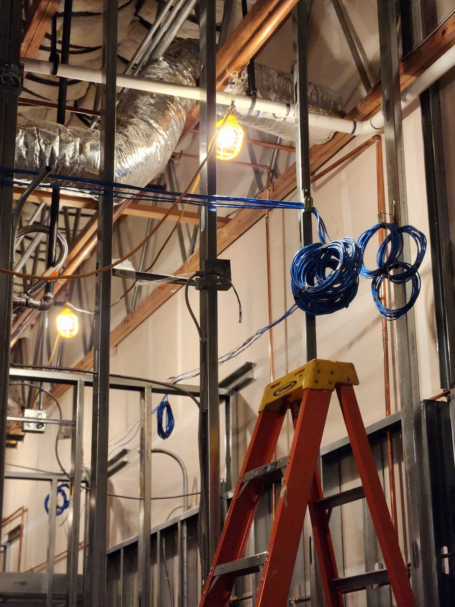 Construction site with metal framing, exposed pipes, electrical wires, and orange ladder.