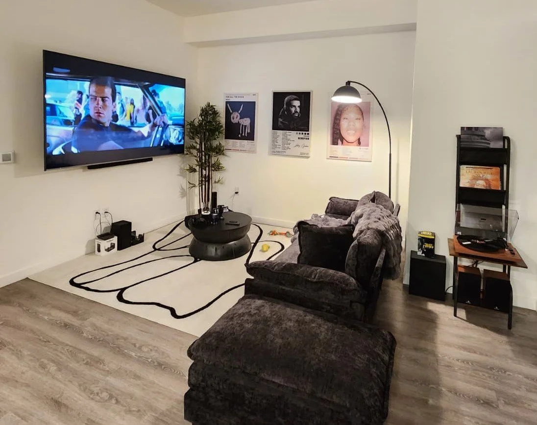 Living room with a large flat-screen TV, a dark plush sectional sofa, a white rug with black lines, a tall floor lamp, and framed posters on the wall.