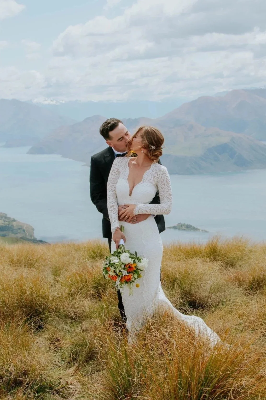 A bride and groom sharing a kiss outdoors on a grassy hill with mountains and a lake in the background.