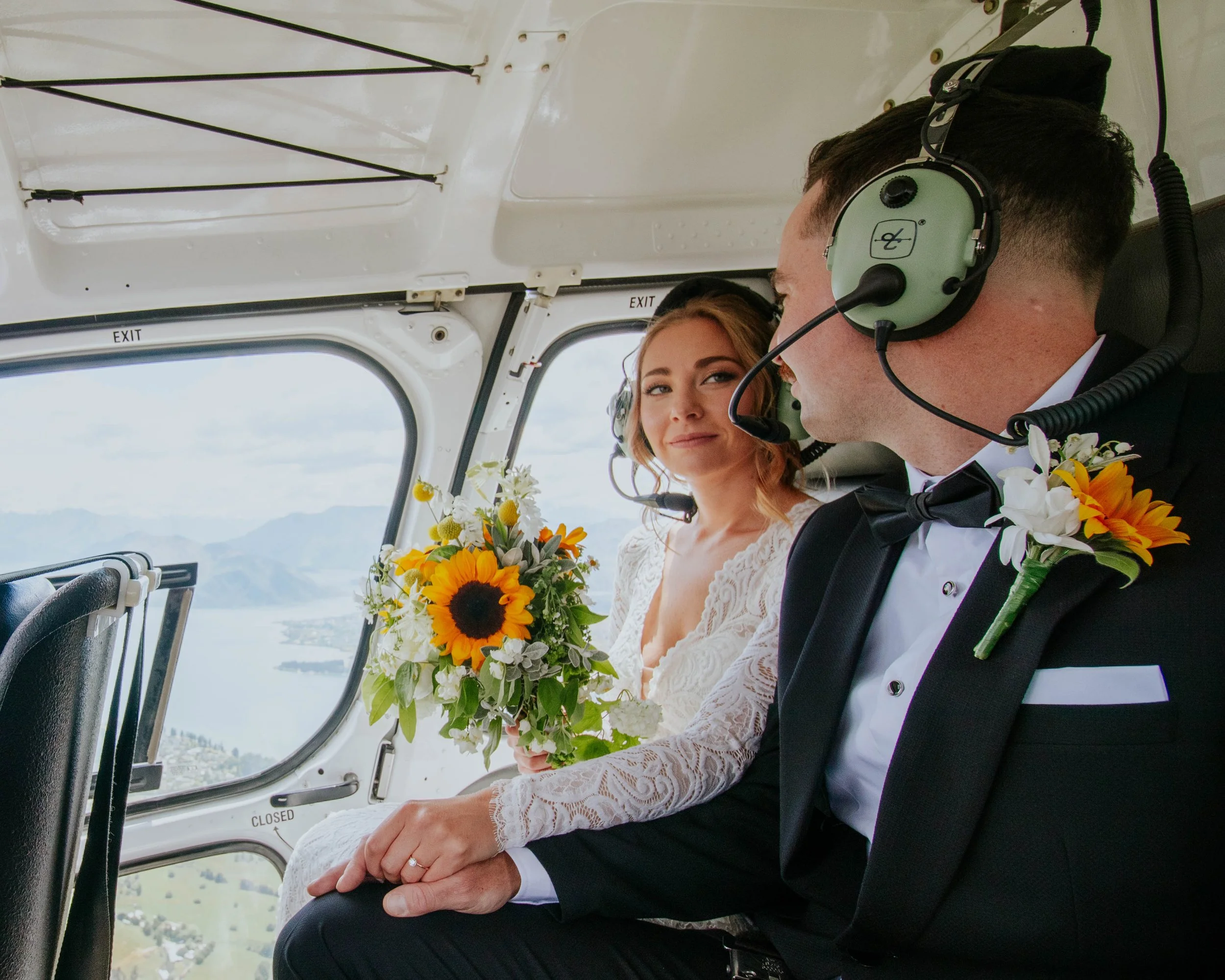 A bride and groom in wedding attire sitting inside a helicopter, holding bouquets with sunflowers and white flowers, during their wedding flight.