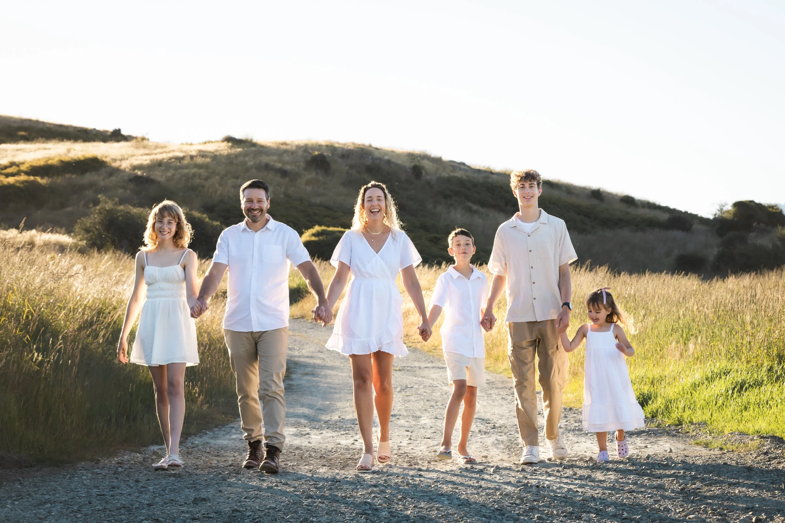 A family of seven walking hand in hand on a dirt trail in a grassy field during sunset, dressed in white clothing.