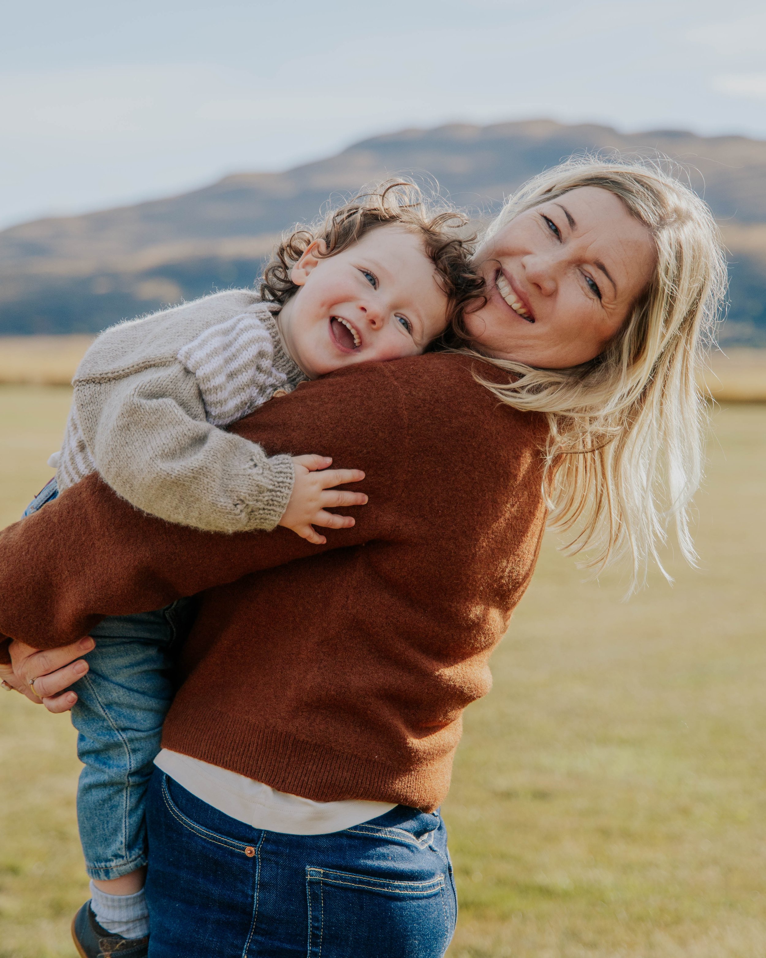 A woman is holding a young child on her back outdoors in a grassy area with mountains in the background. The woman is smiling, and the child appears happy and laughing.