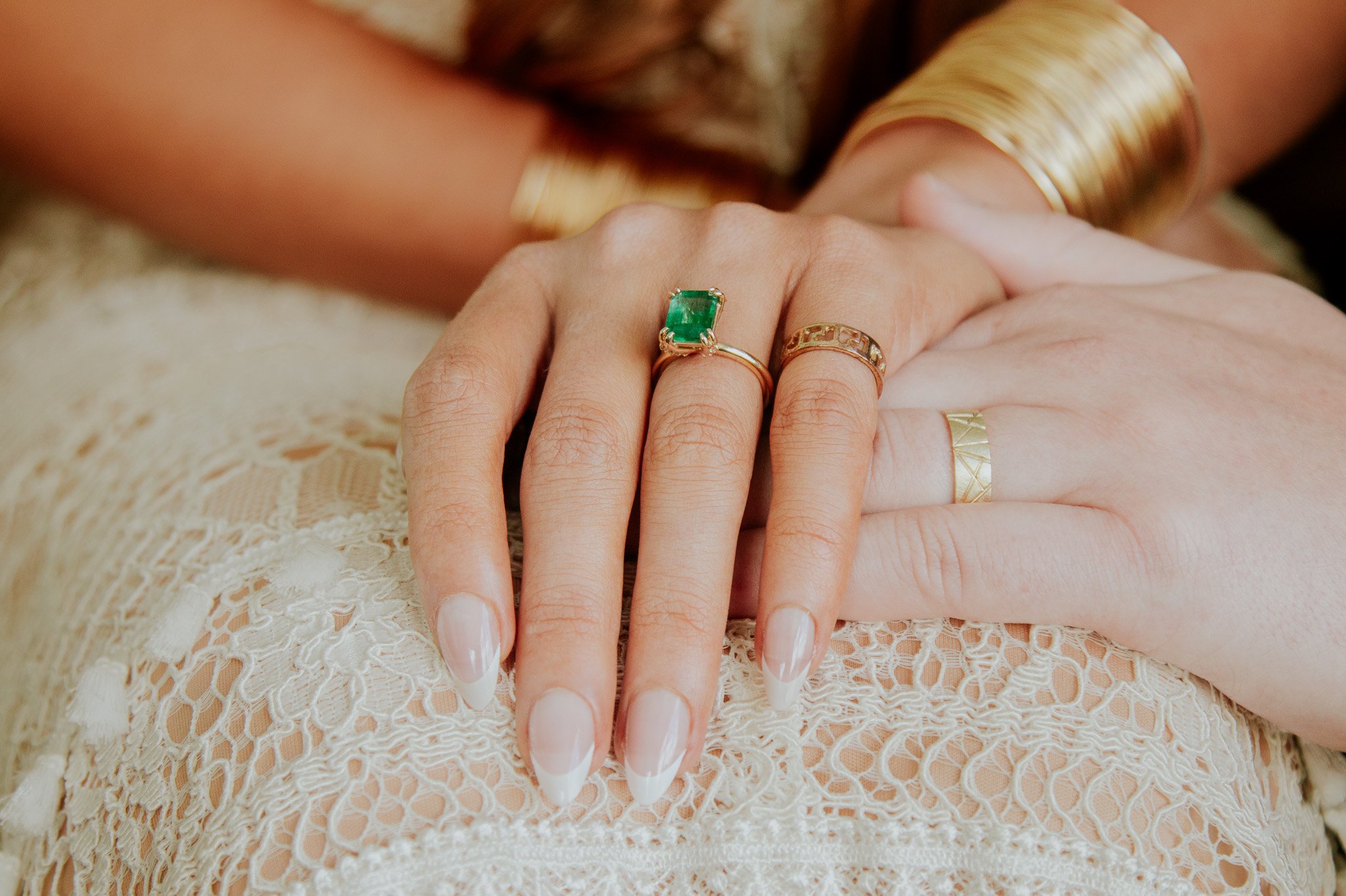 Close-up of two hands with rings, one with an emerald ring, resting on lace fabric. Gold bangles are visible on the wrist.