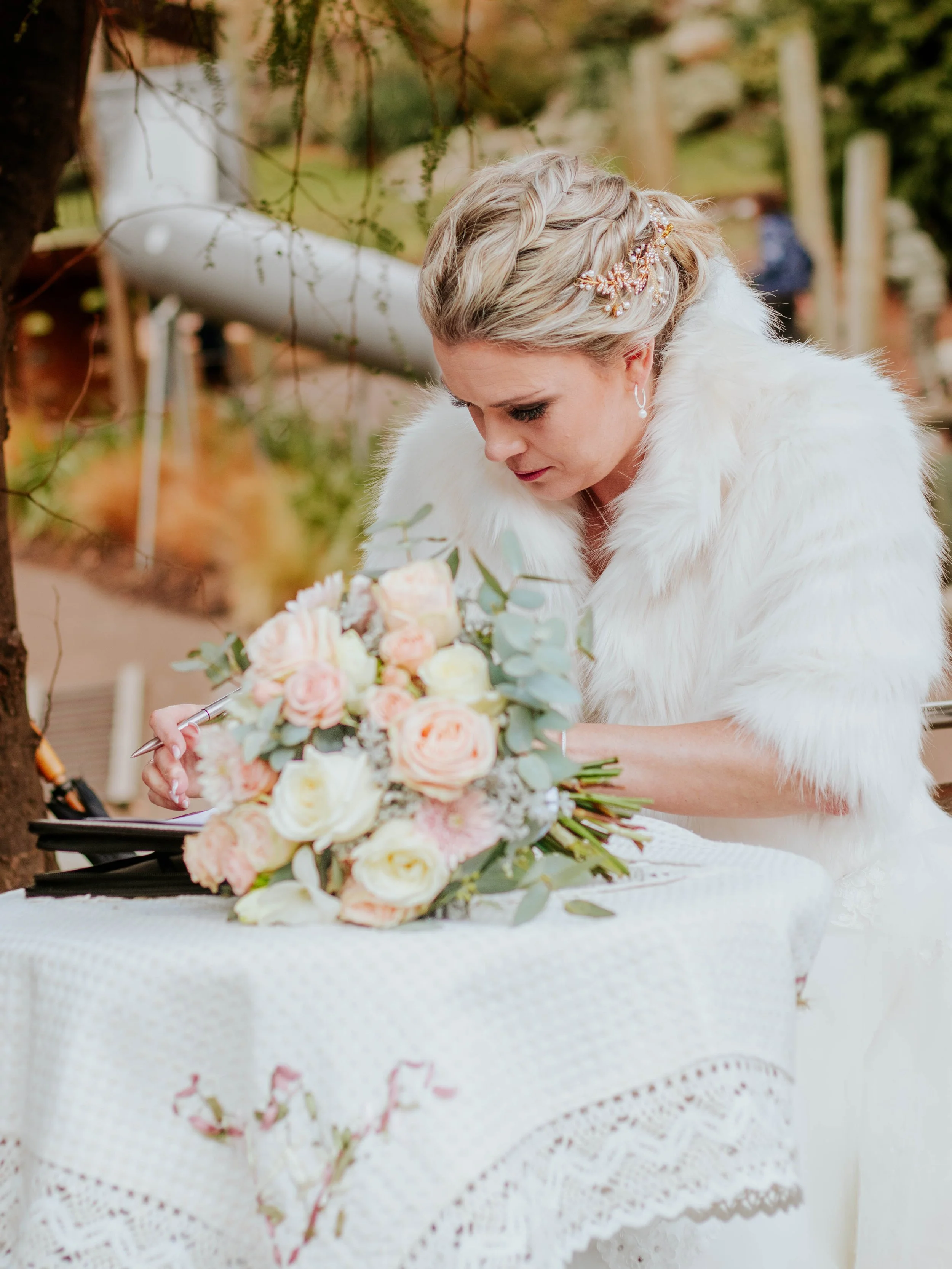 Bride signing a marriage register with a bouquet of pink and white roses on the table in front of her.