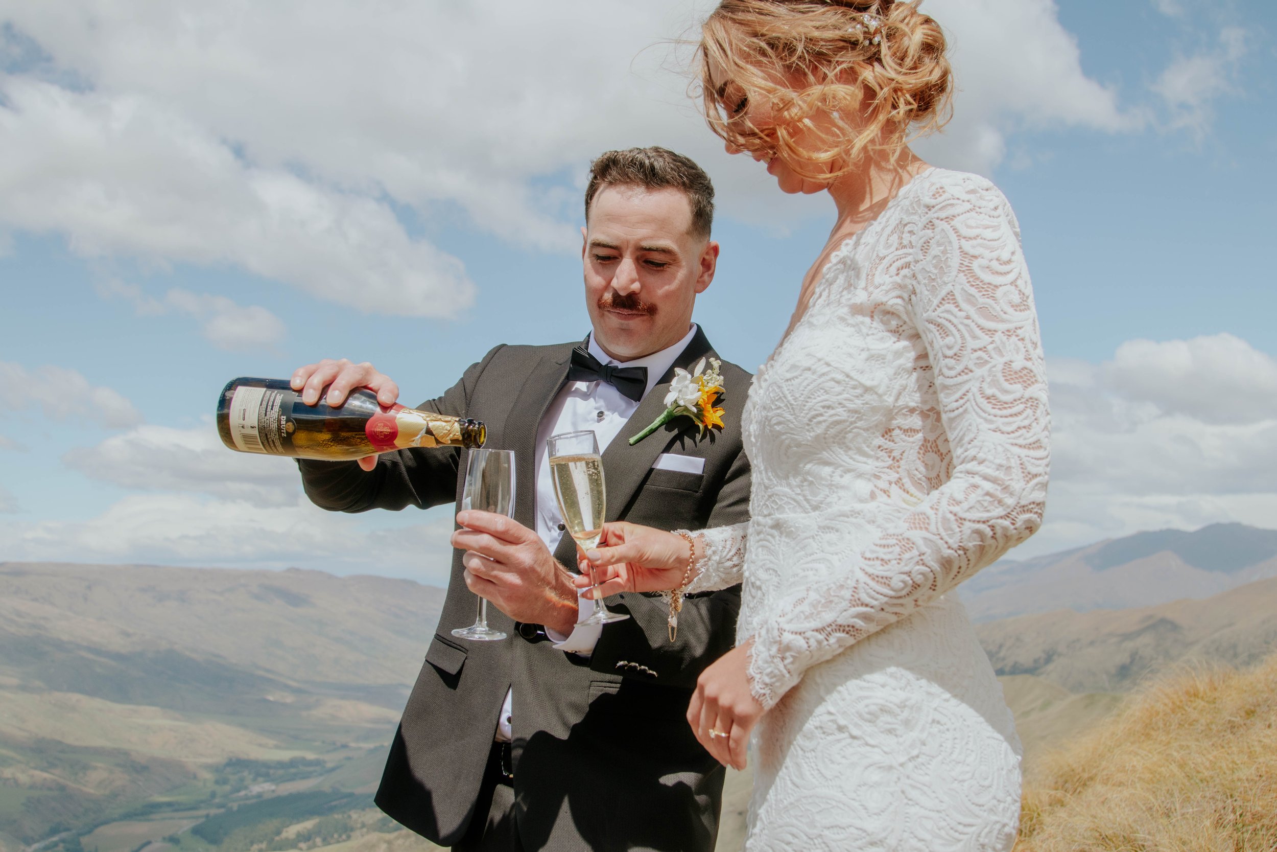 A couple dressed in wedding attire pouring champagne into glasses outdoors under a blue sky with clouds, mountains in the background.