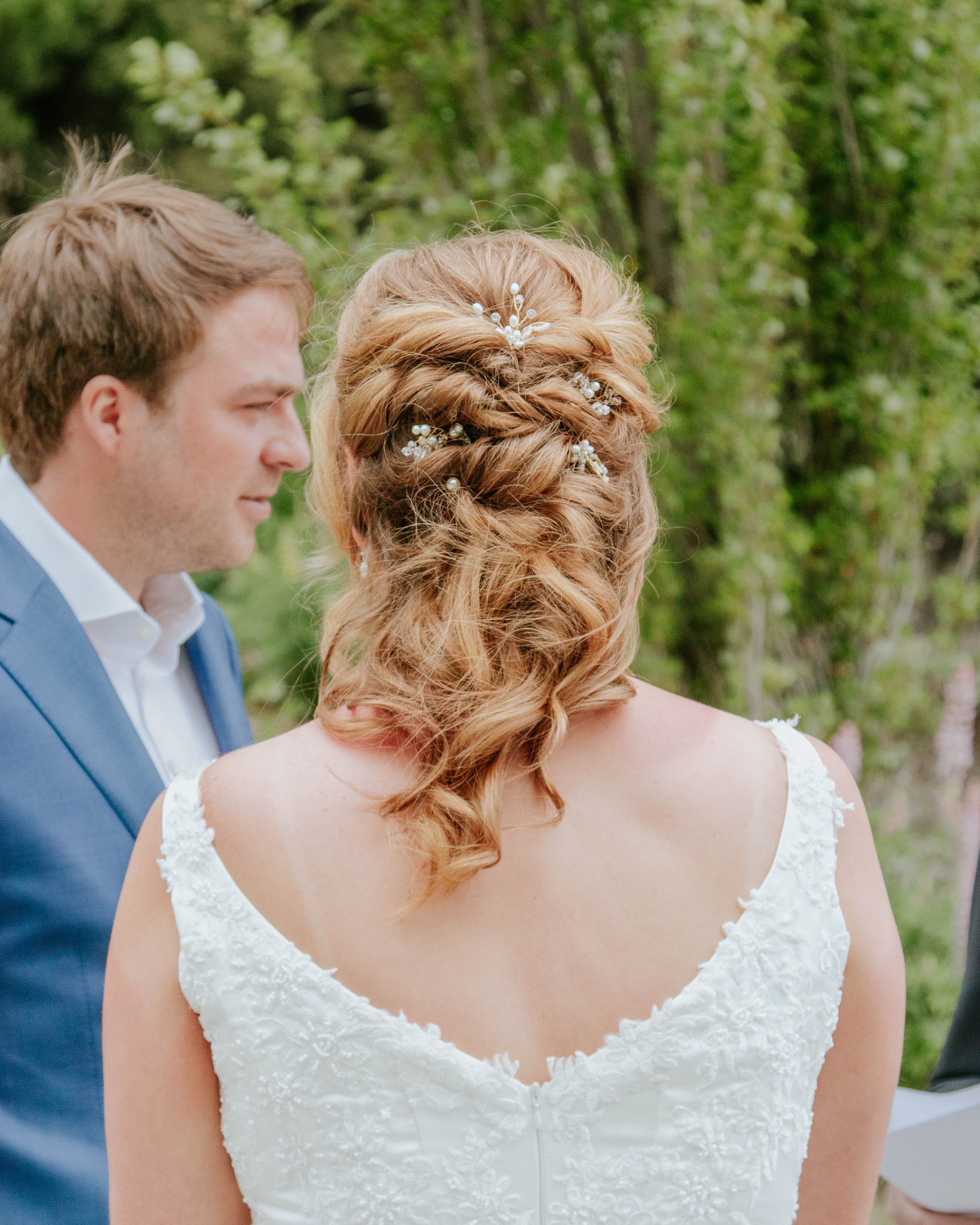 Bride and groom standing outdoors during wedding ceremony, with the bride's back to the camera, showing her curly red hair adorned with pearl and floral hairpins, and wearing a lace wedding gown, while the groom is in a blue suit, facing her.