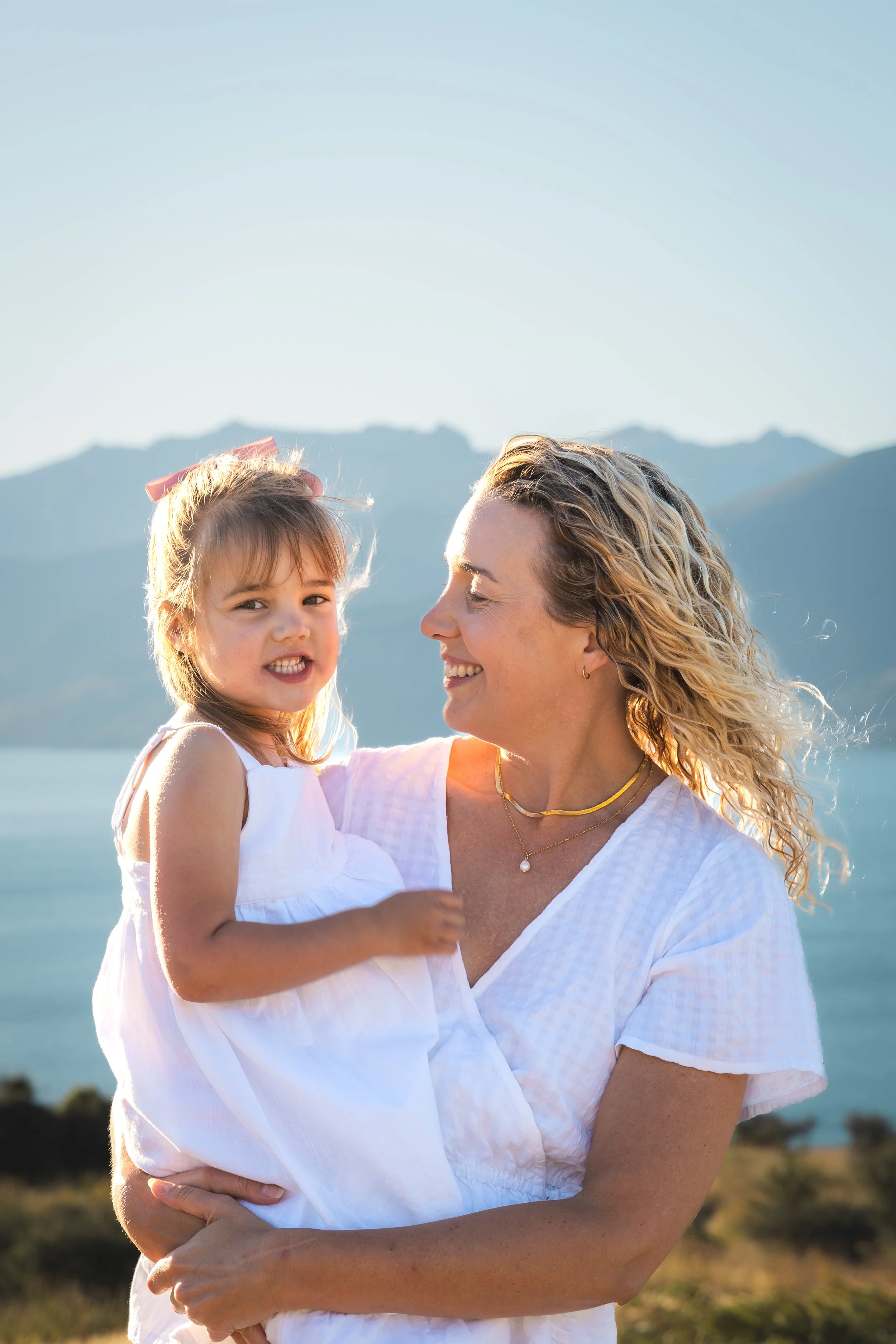 A woman holding a young girl outdoors with mountains and a body of water in the background, both smiling.