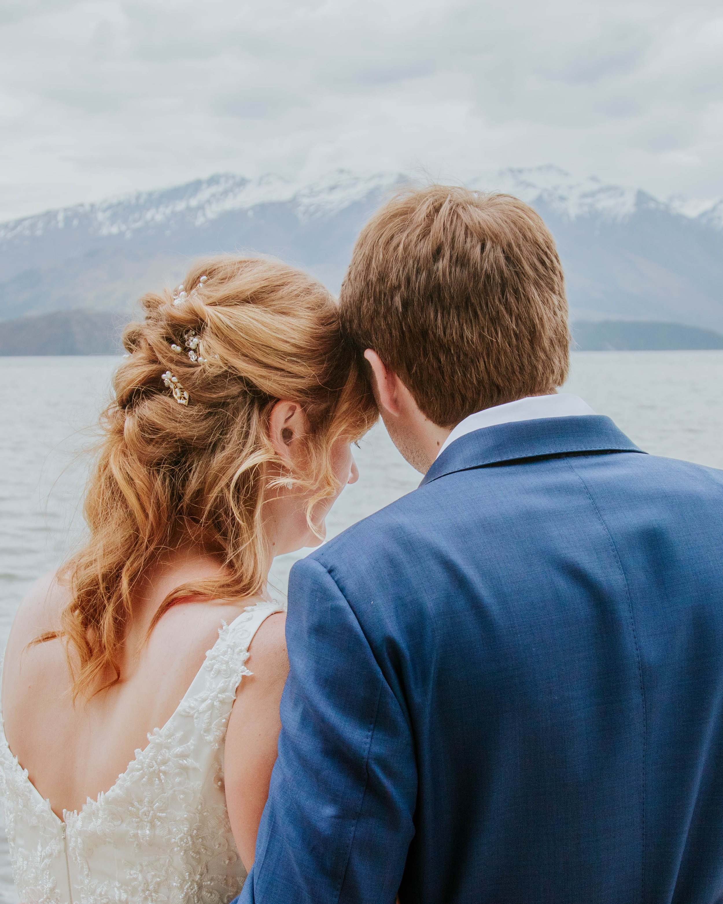 A couple dressed in wedding attire embracing near a lake with snow-capped mountains in the background.