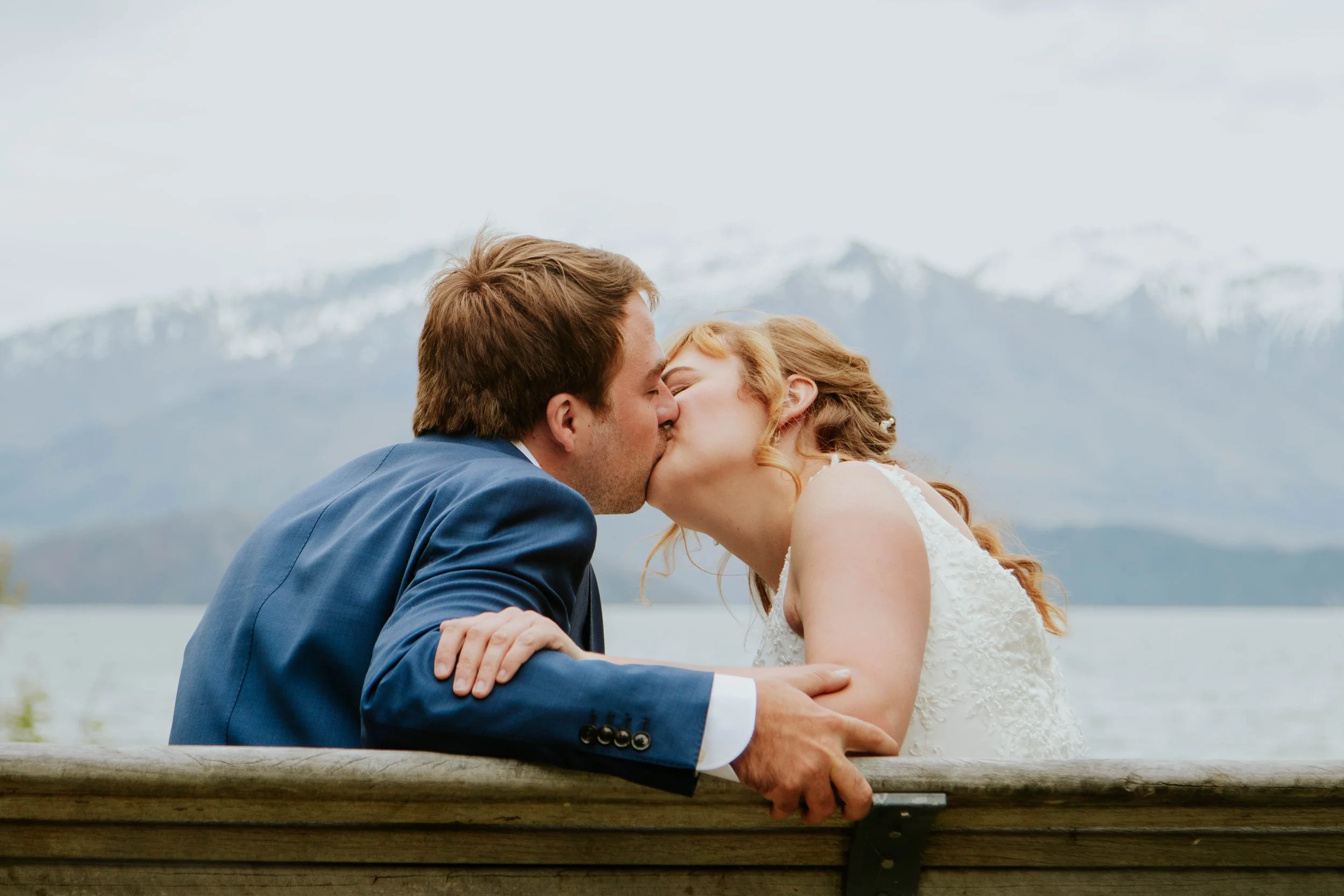 A couple dressed in wedding attire kissing by a lake with snow-capped mountains in the background.