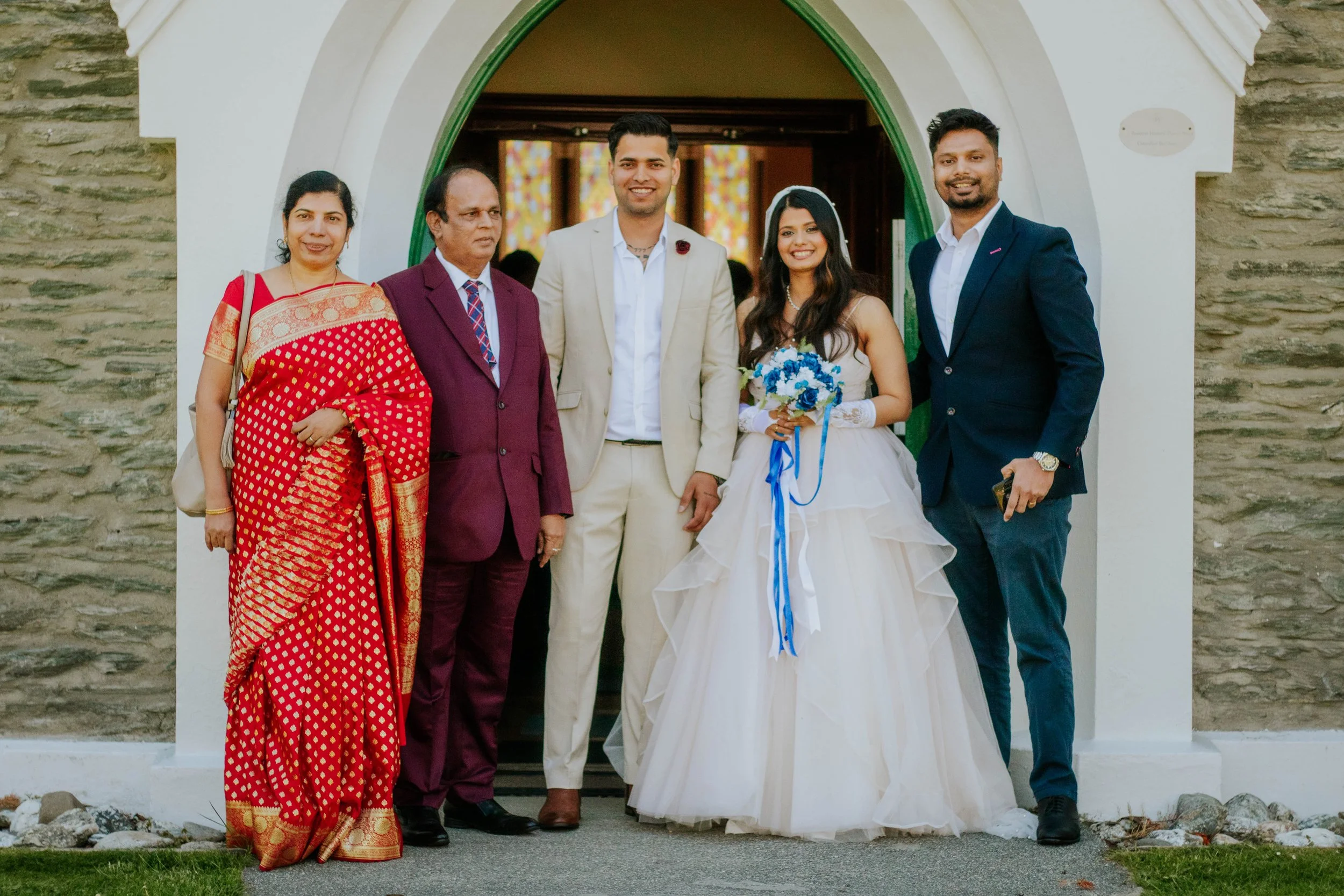 Group of six people standing in front of a church entrance, including a bride in a white wedding dress holding a bouquet, a groom in a light-colored suit, and four others in formal and traditional attire.