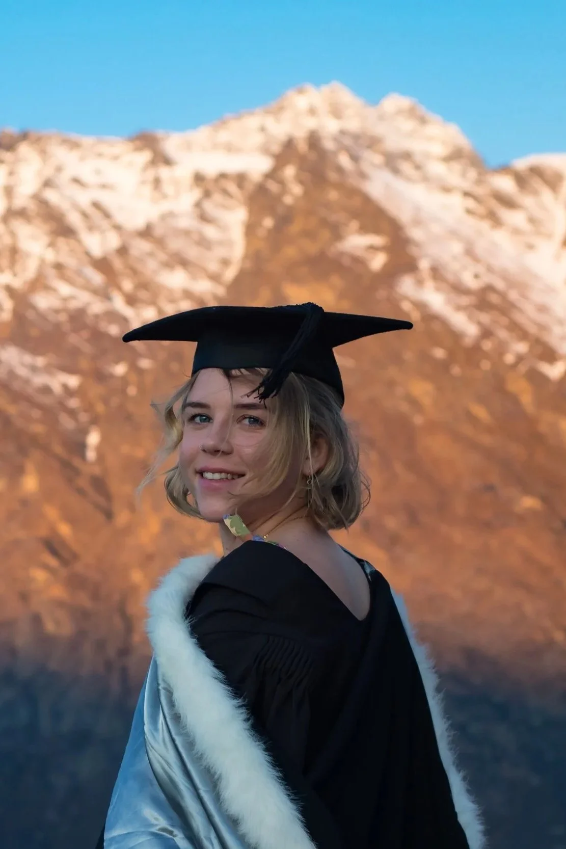 Young woman in a graduation cap and gown with a snowy mountain in the background.