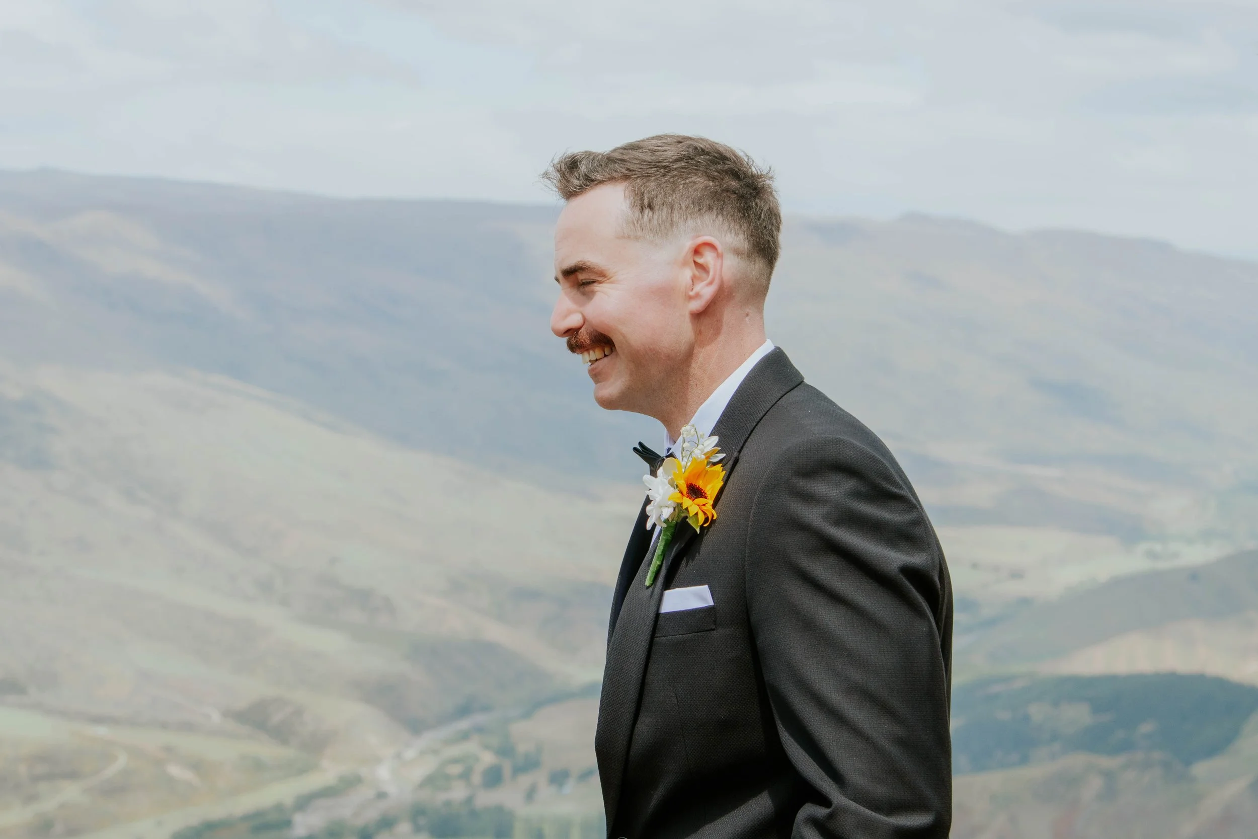 Man in a black suit with a white shirt and a floral boutonniere, smiling outdoors with mountains in the background.
