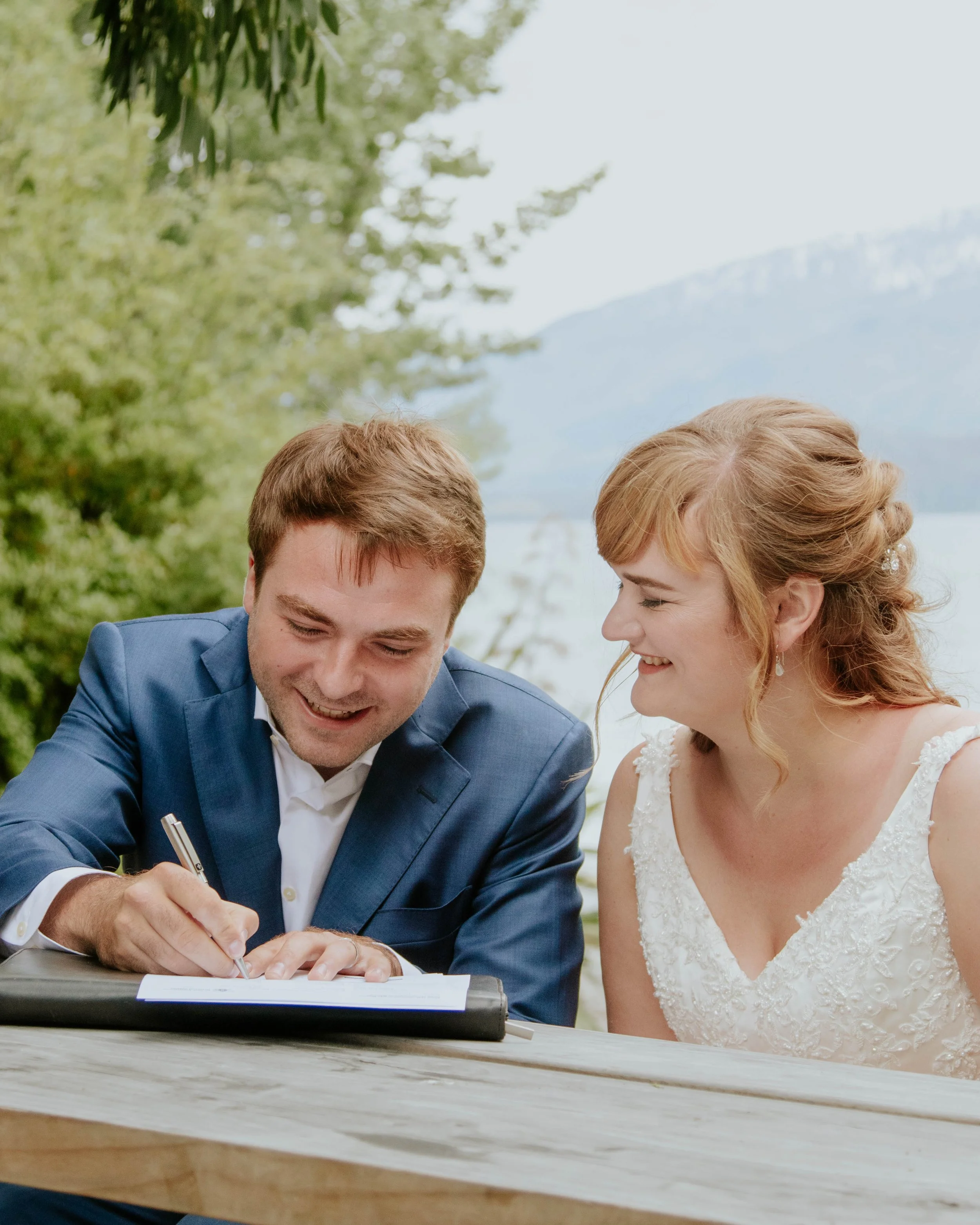 A couple is sitting at a wooden table outdoors, with the man signing a document and both smiling, in a scenic natural setting with trees, mountains, and a lake in the background.