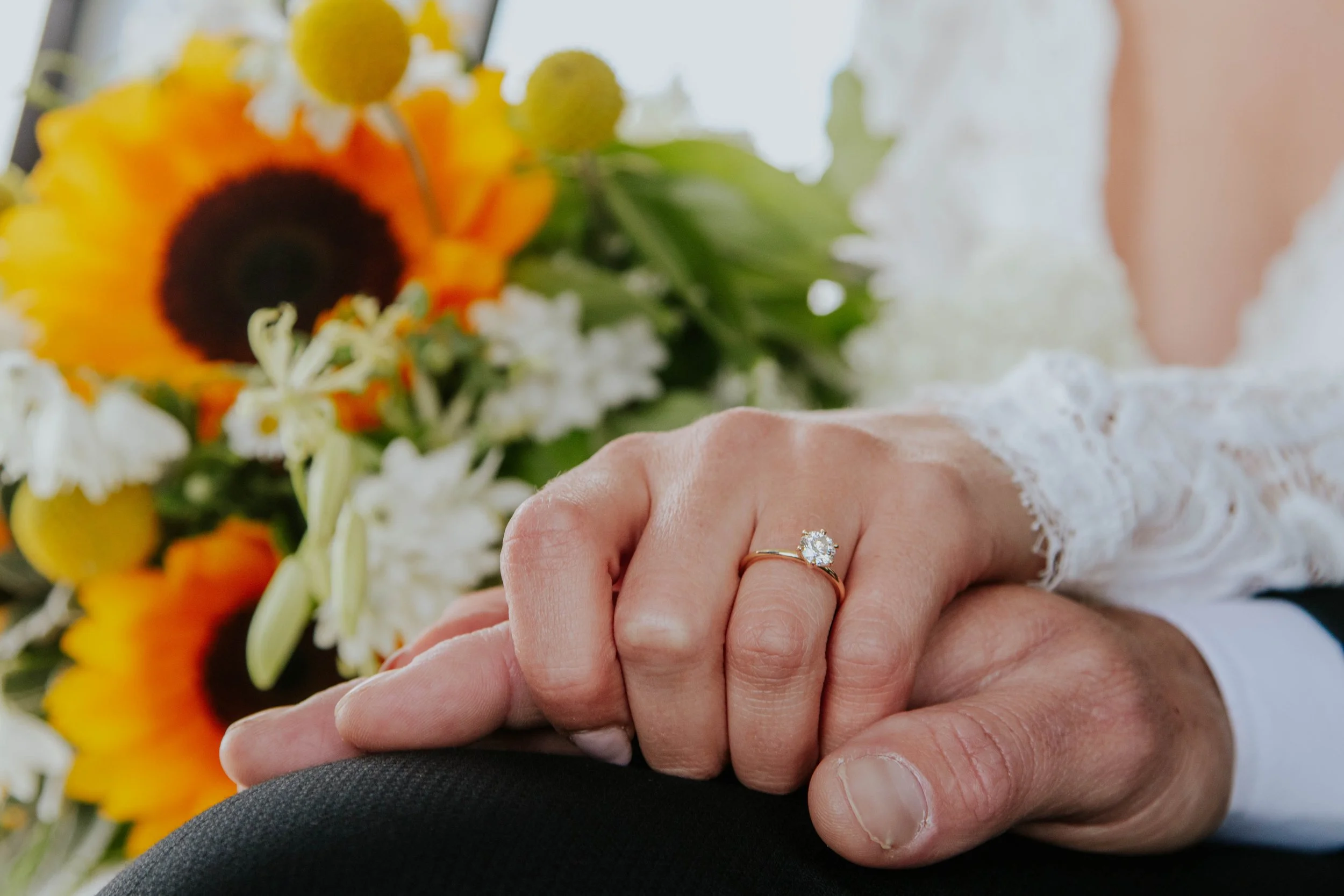 Close-up of a woman’s hand with an engagement ring resting on a man’s hand, with a bouquet of colorful flowers including sunflowers and white blooms in the background.