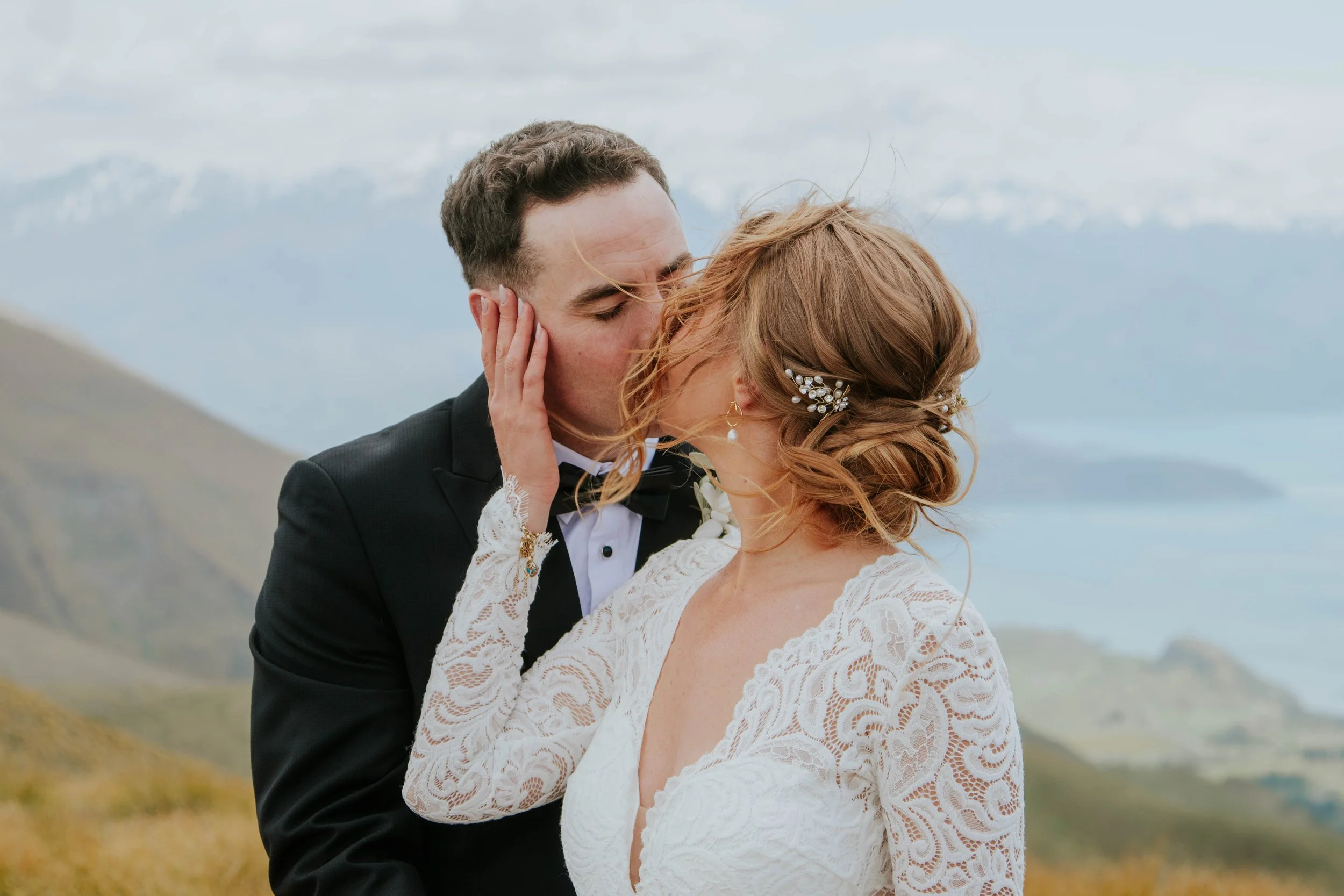 A bride and groom sharing a kiss outdoors on a hill with mountains and cloudy sky in the background. The bride has red hair with a decorative hairpiece and wears a white lace dress. The groom has dark hair and is dressed in a black tuxedo.