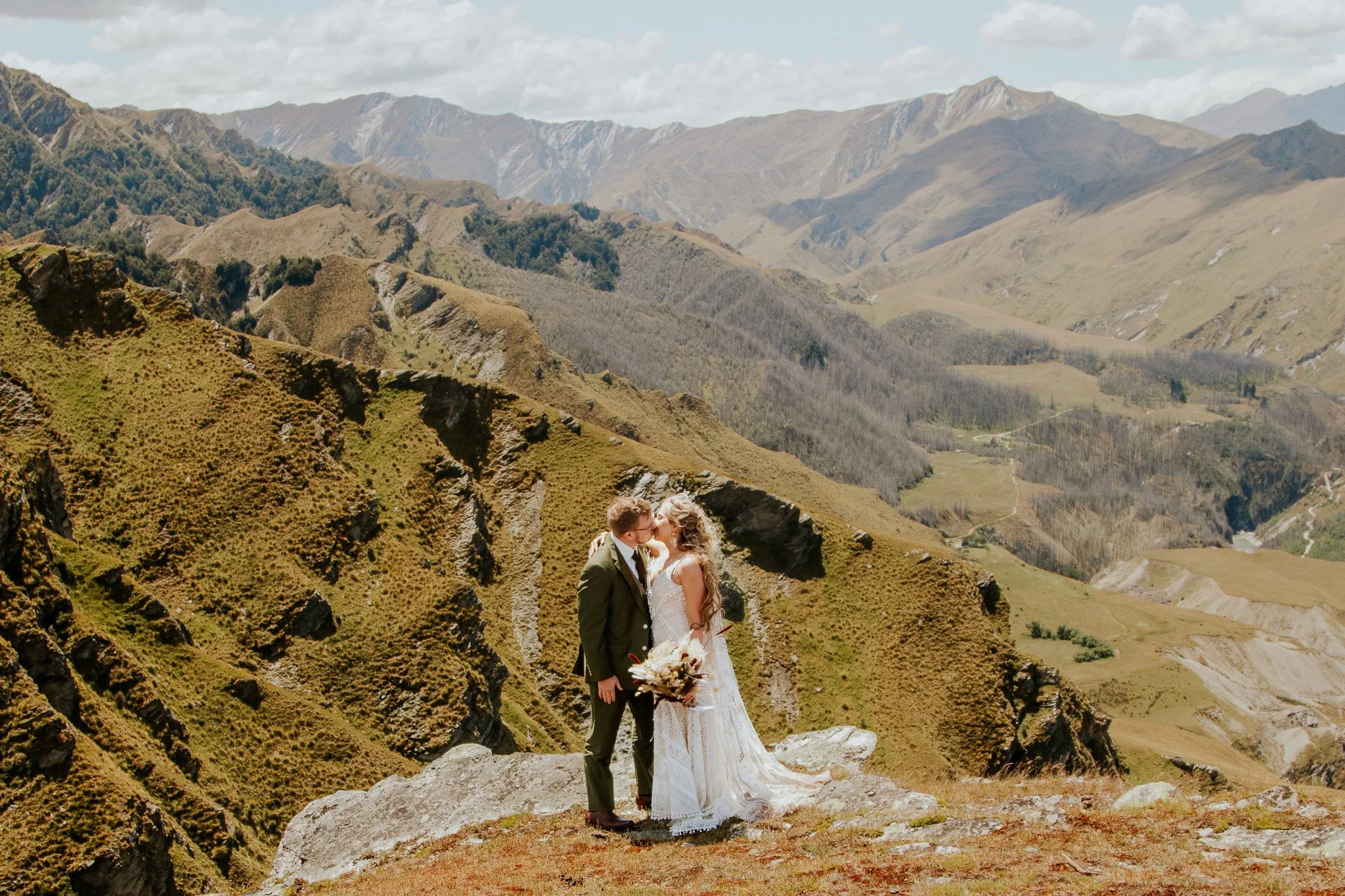 A couple in wedding attire sharing a kiss on a rocky outcrop in a mountainous landscape with green hills and mountains in the background.