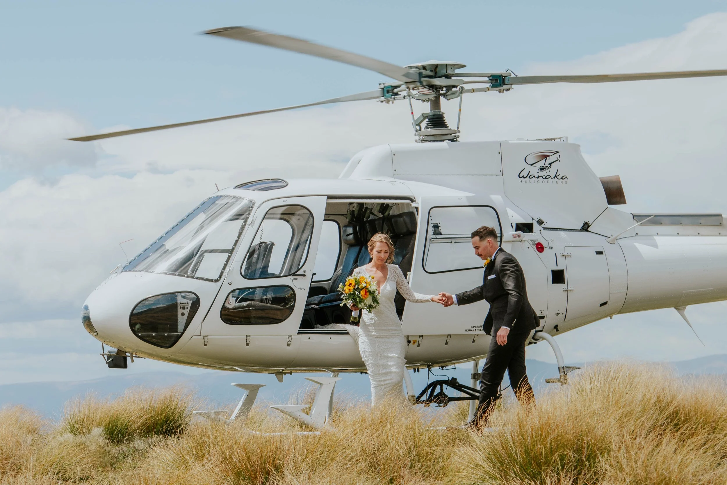 A bride and groom holding hands in front of a white helicopter on a grassy field, with a cloudy sky in the background.