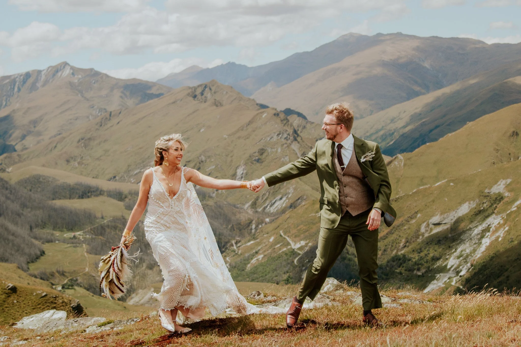 A bride and groom holding hands on a grassy hillside with mountainous terrain in the background. The bride wears a white lace wedding dress and the groom is dressed in a green suit with brown shoes.