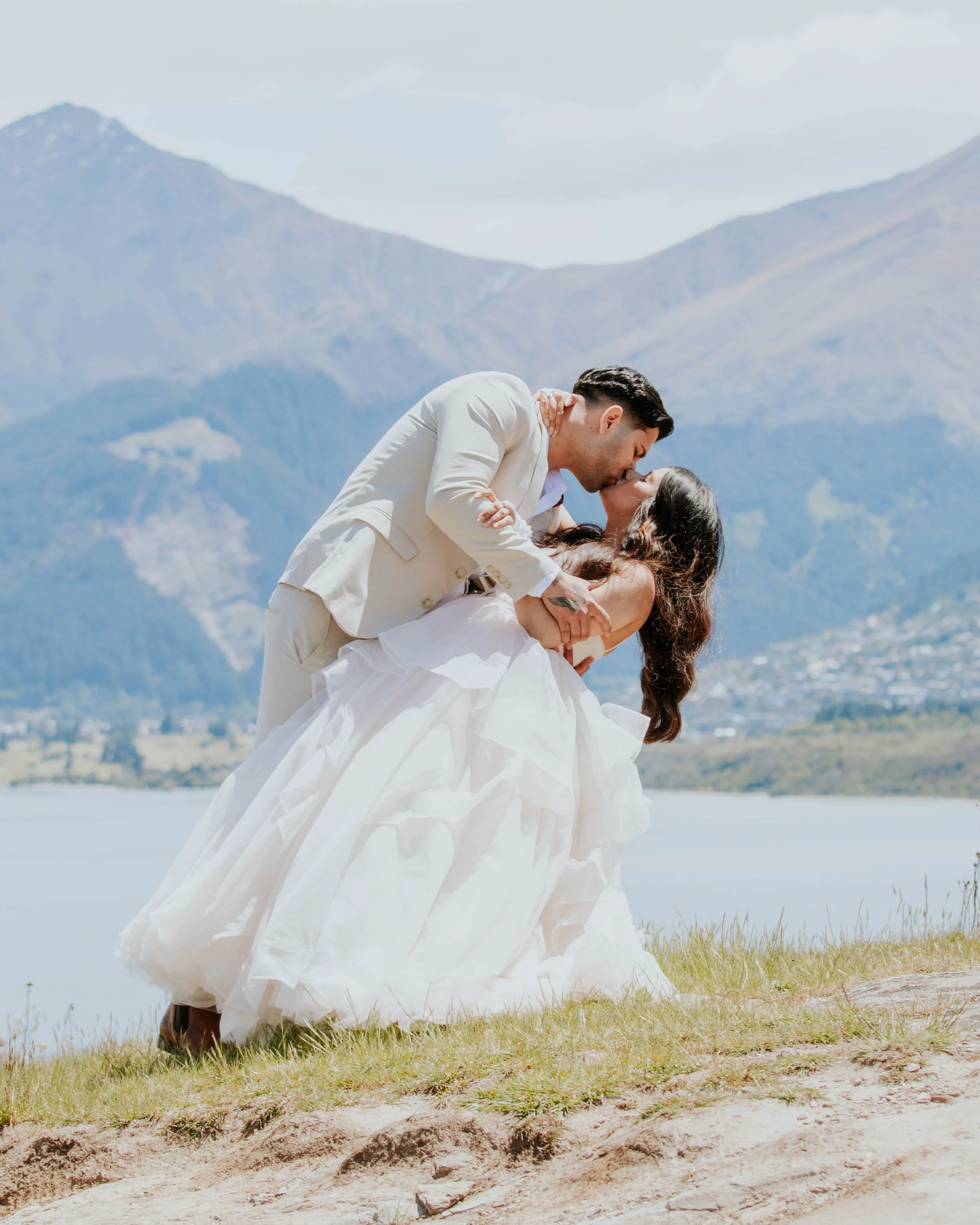 A couple in wedding attire sharing a kiss outdoors near a lake with mountains in the background.