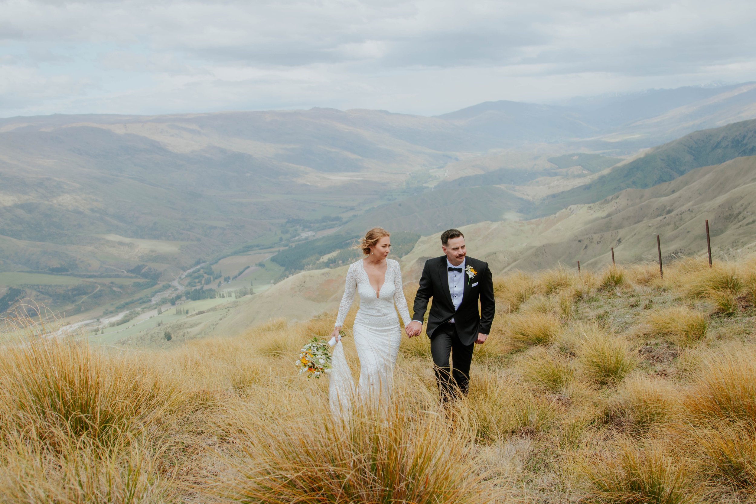 A bride and groom walking hand in hand through a grassy hillside with mountains and cloudy sky in background.