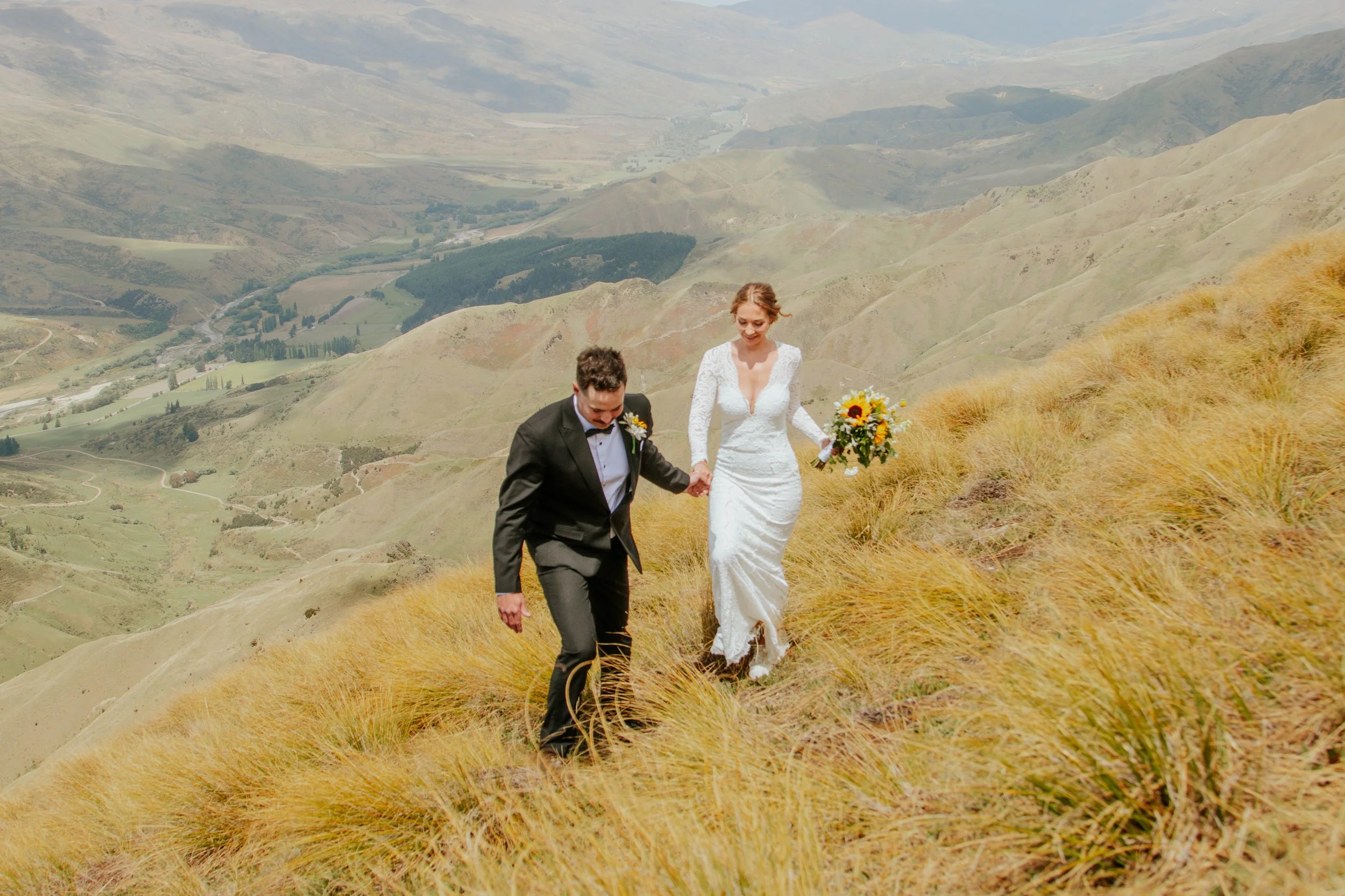 A bride and groom holding hands and walking on a grassy hillside with a scenic valley and rolling hills in the background, during a wedding photoshoot.