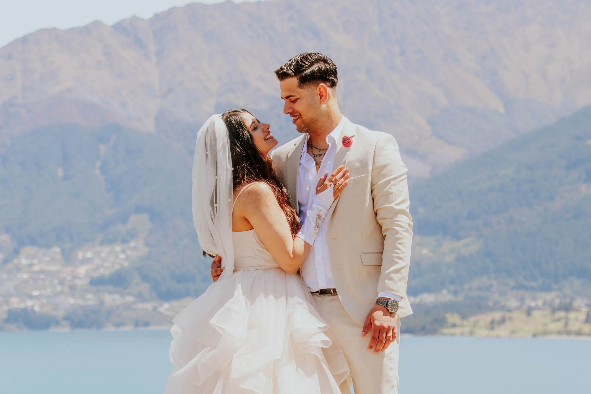 A bride and groom sharing a joyful moment outdoors near a lake with mountains in the background.