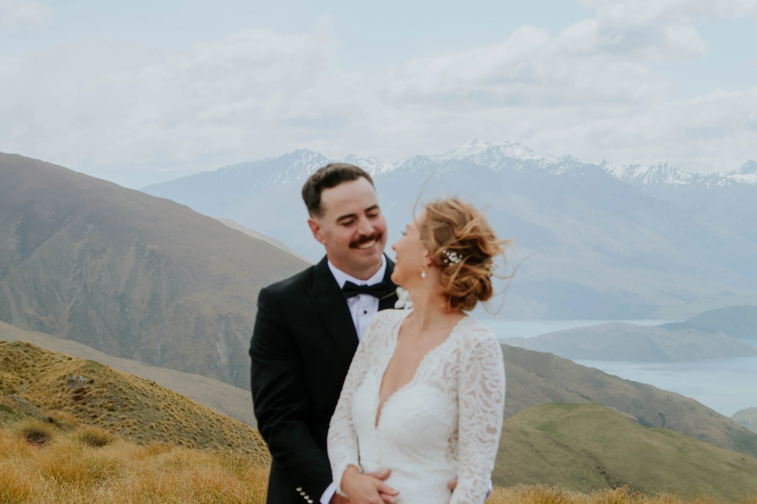 A newlywed couple smiling and looking at each other outdoors on a mountainous landscape with cloudy sky.