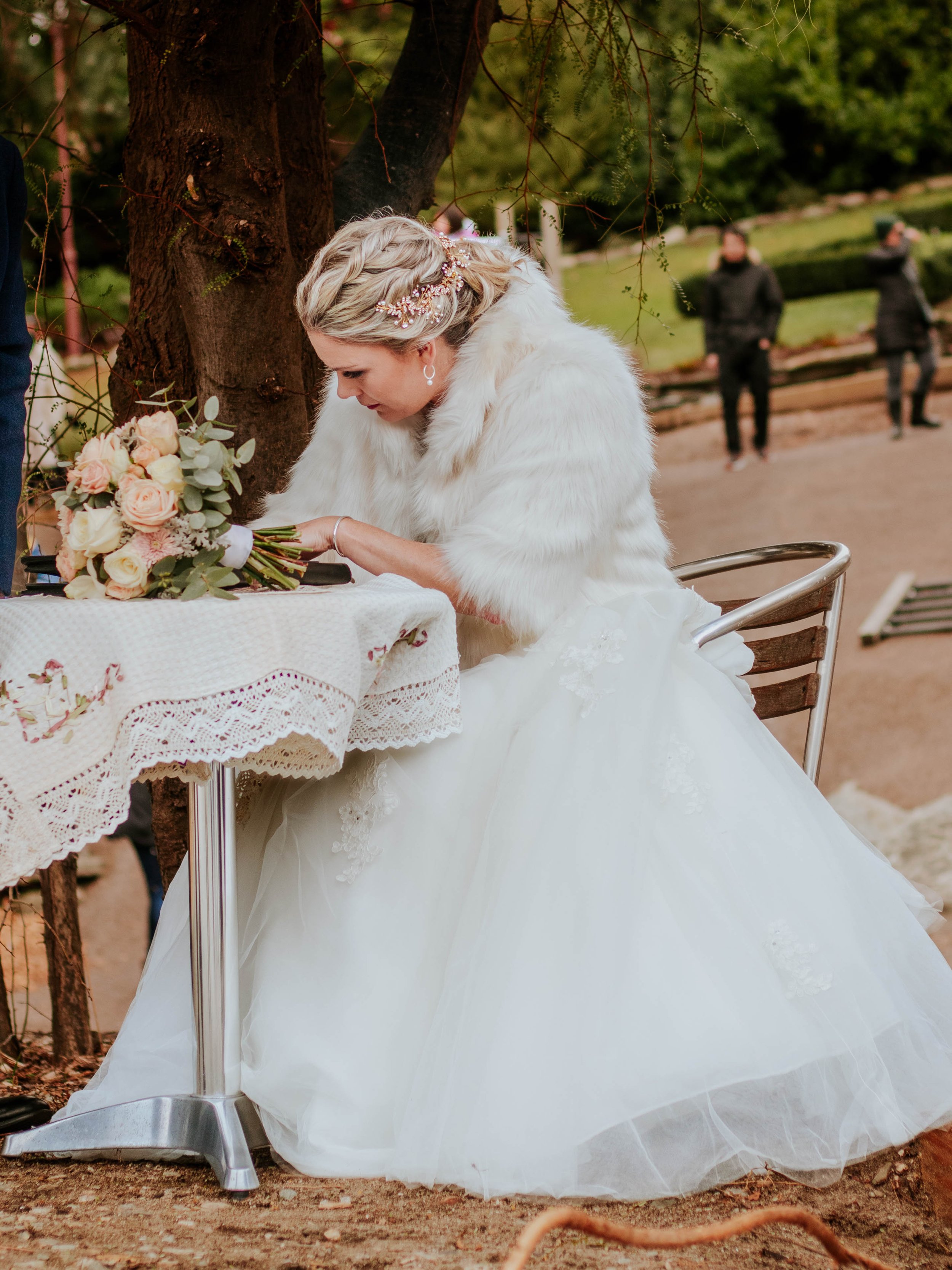 A bride in a white wedding gown and furry white shawl, sitting at an outdoor table under a tree, signing a document with a bouquet of pale pink and cream roses on the table.