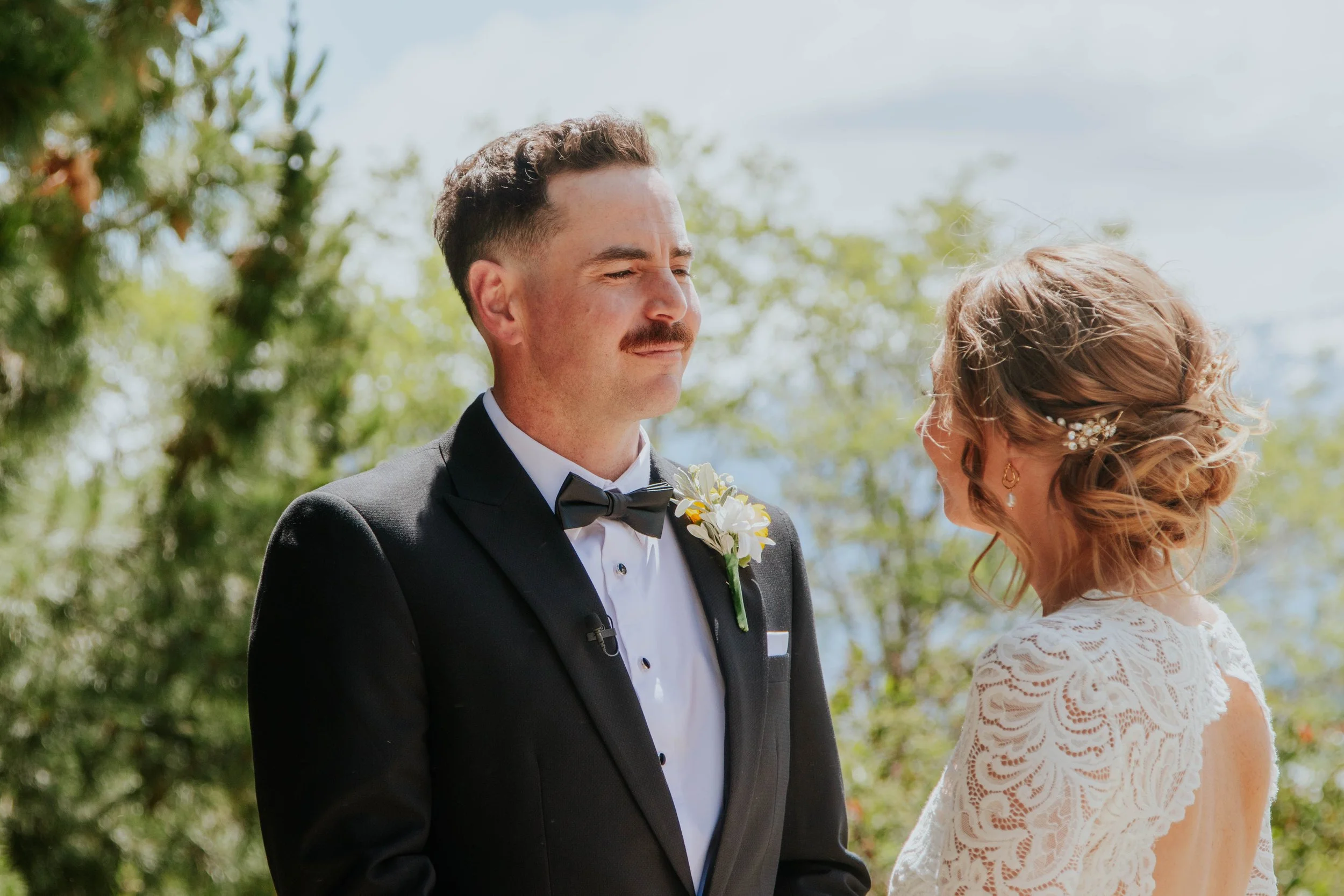 A groom in a black tuxedo with a bow tie and boutonniere stands outdoors during a wedding ceremony, smiling at a bride with styled hair, earrings, and a lace dress.