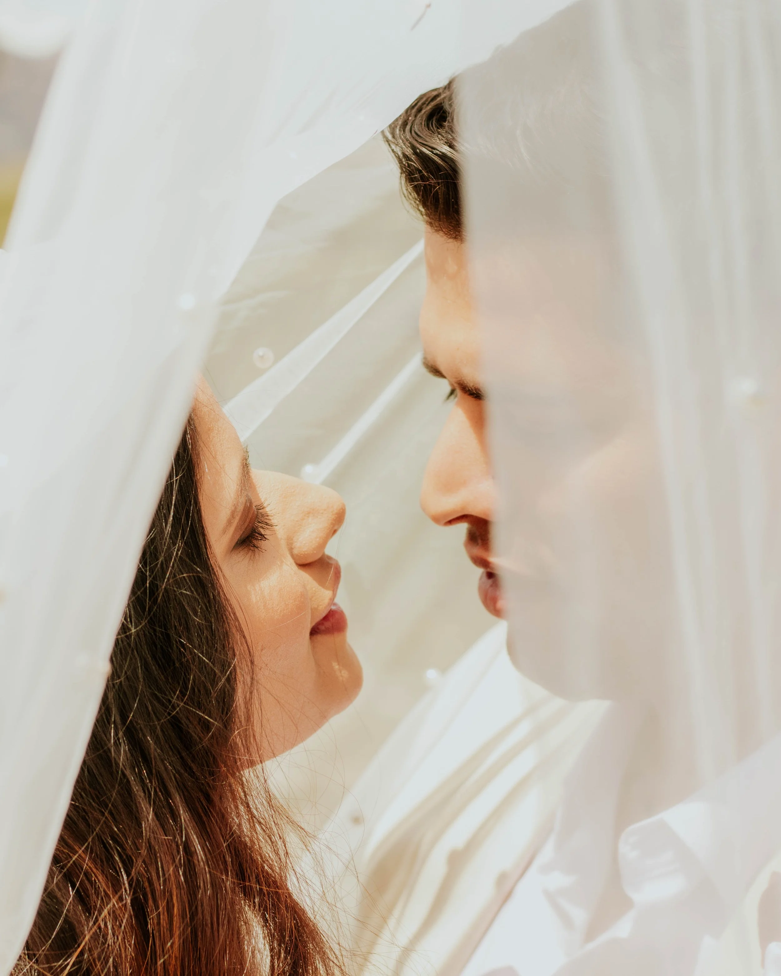A couple under a sheer white veil, nose to nose, close-up shot.
