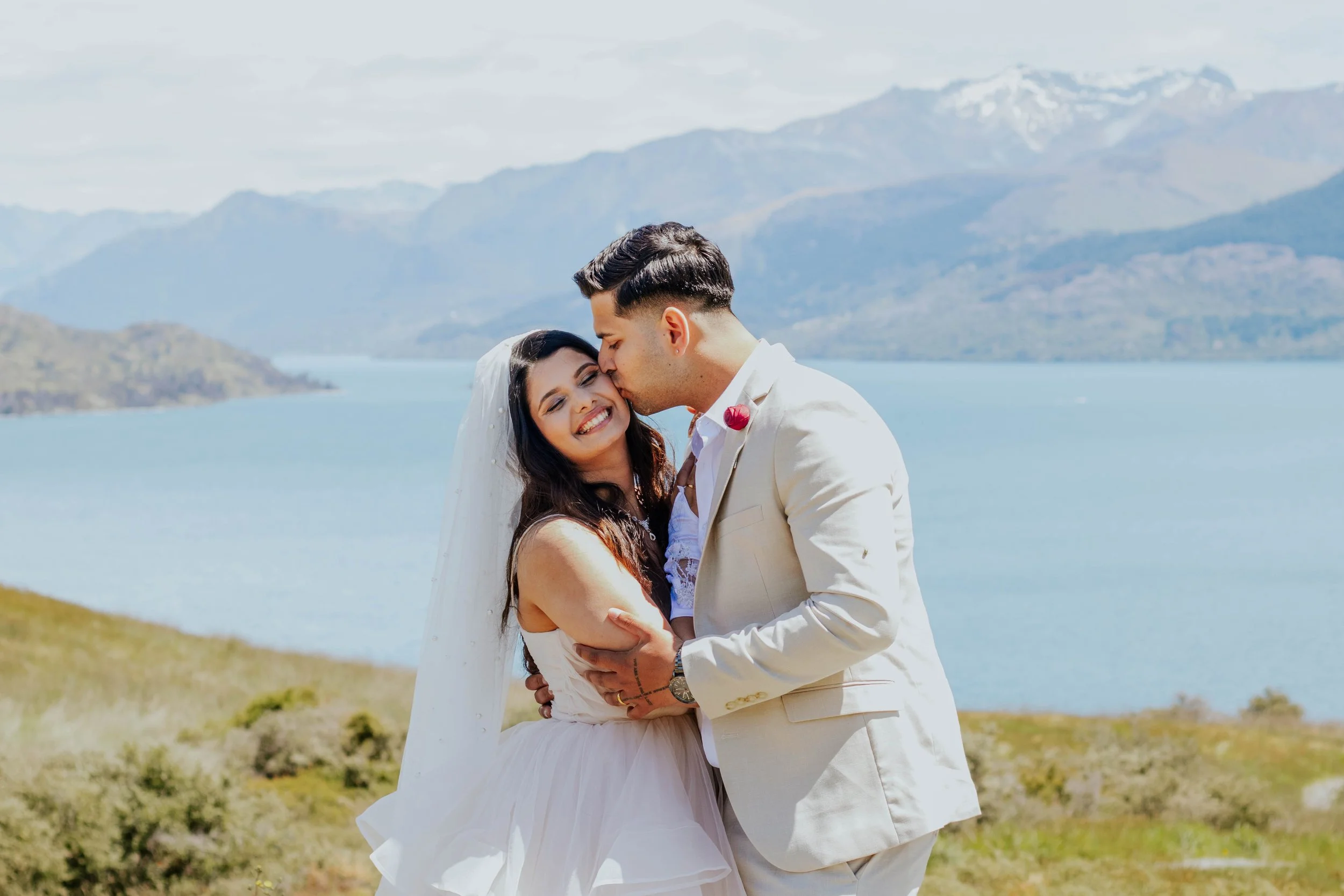 A bride and groom share a kiss and embrace outdoors near a lake with mountains in the background, under a partly cloudy sky.