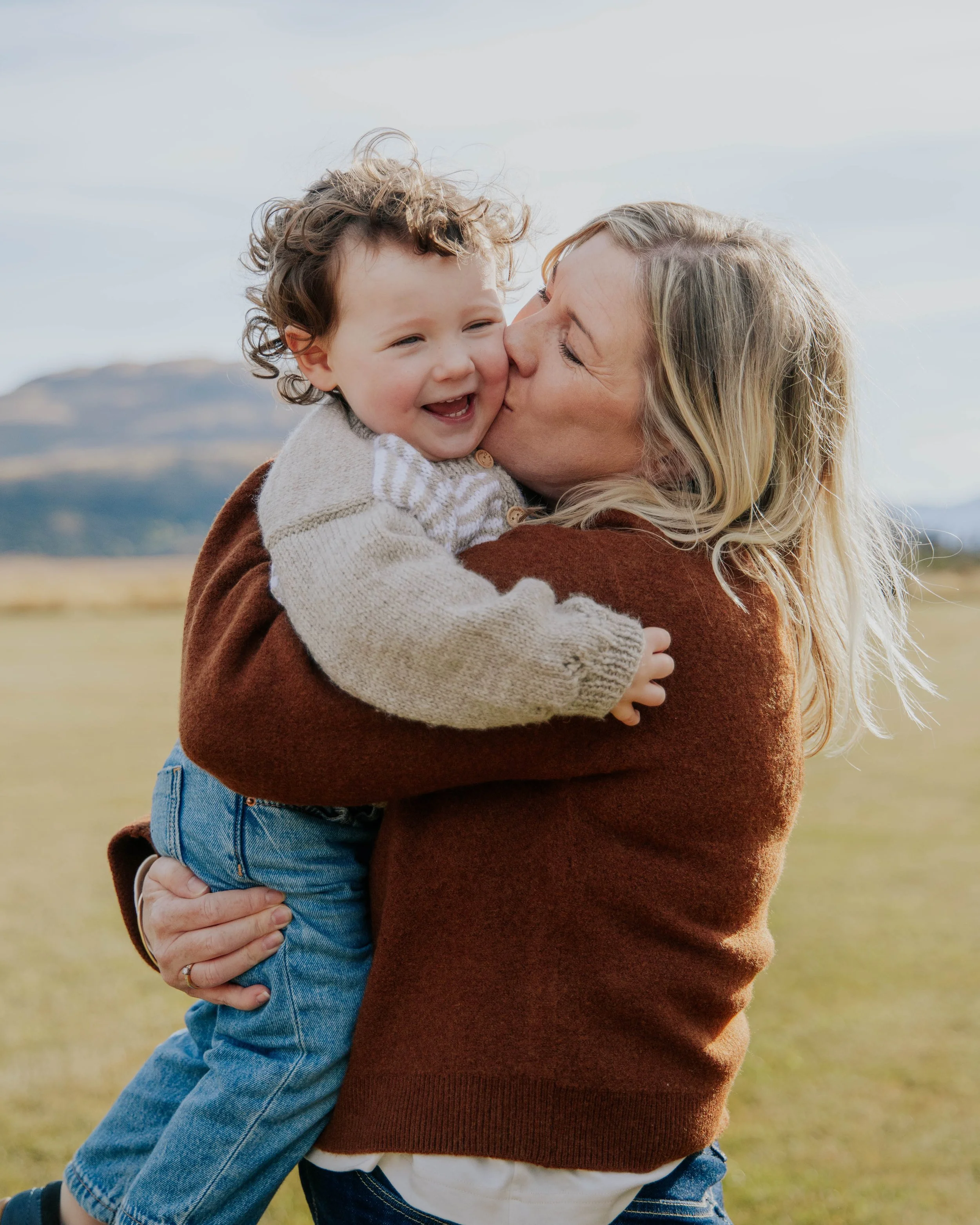 A woman with blonde hair kisses a smiling young boy with curly hair. They are outdoors in a field with a mountain in the background.