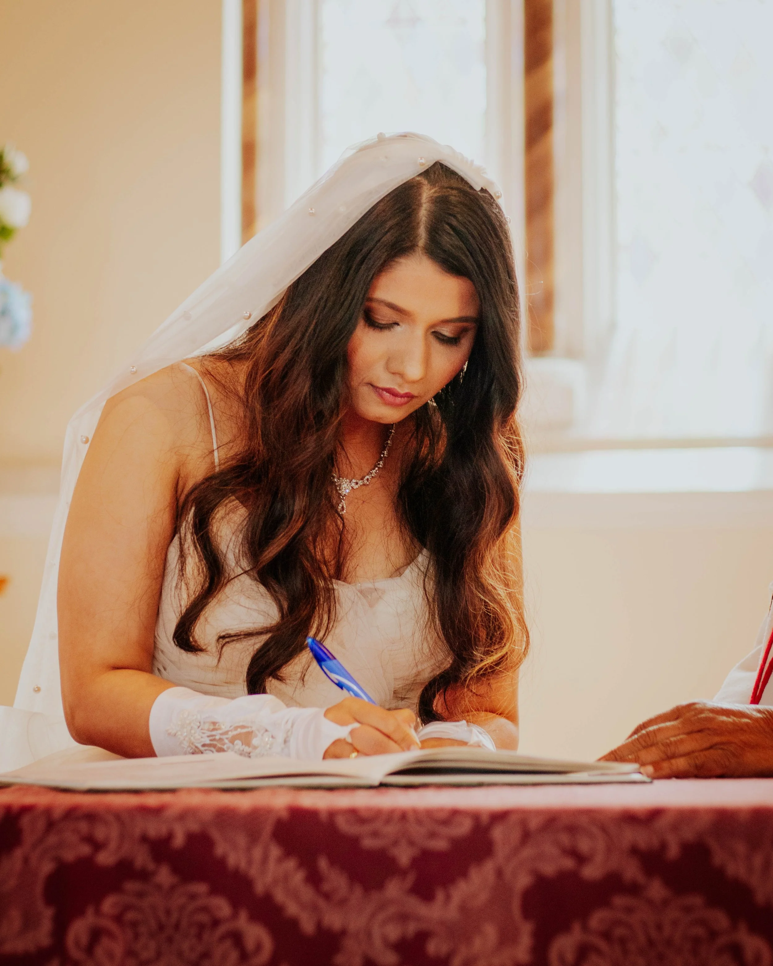 A bride with long dark hair and a white wedding dress is signing a document with a blue pen. She is wearing jewelry and a veil, and is seated at a table covered with a red patterned cloth. A person's hand is visible on the right side.