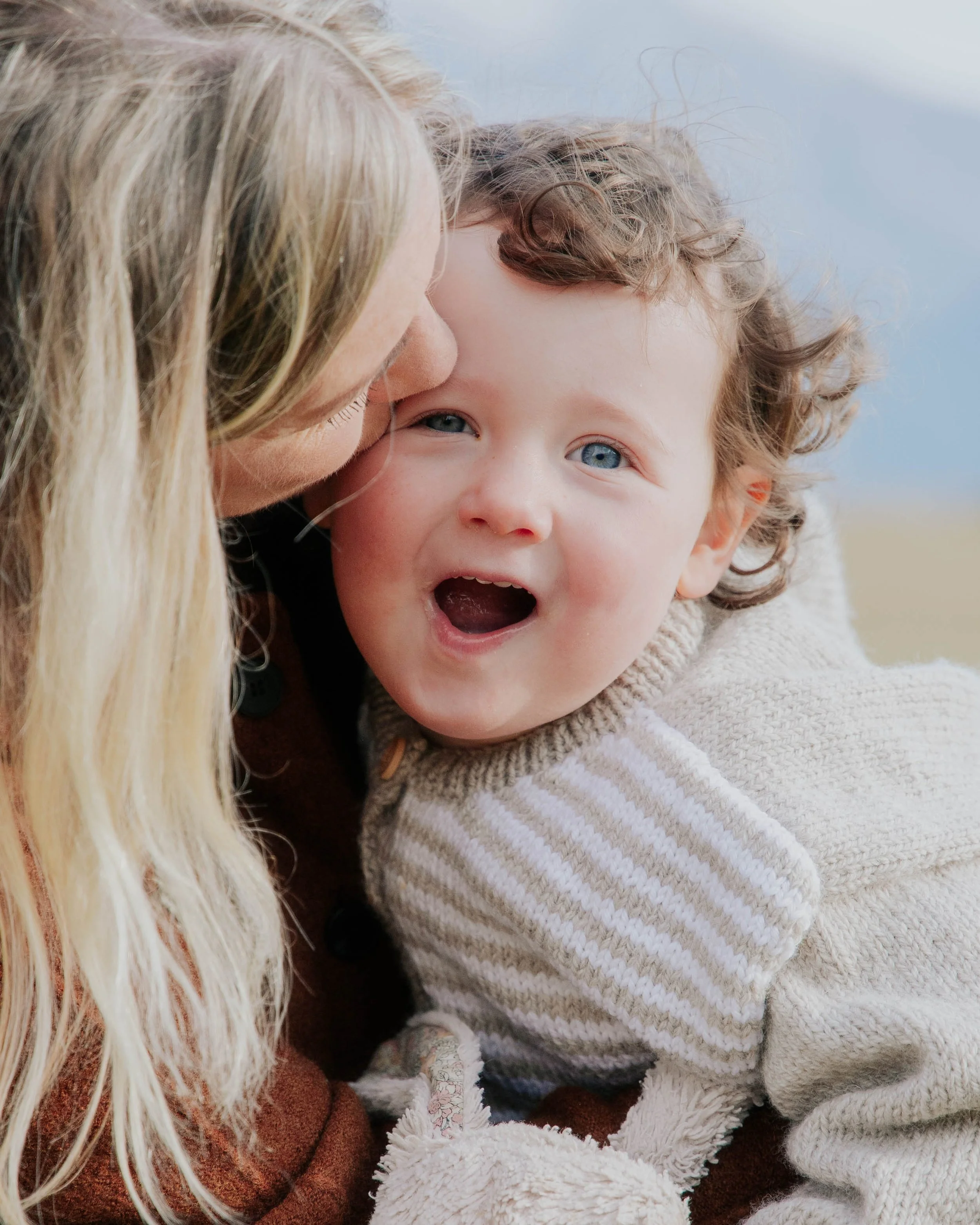 A woman with blonde hair and a young child with curly hair embracing outdoors during daytime.