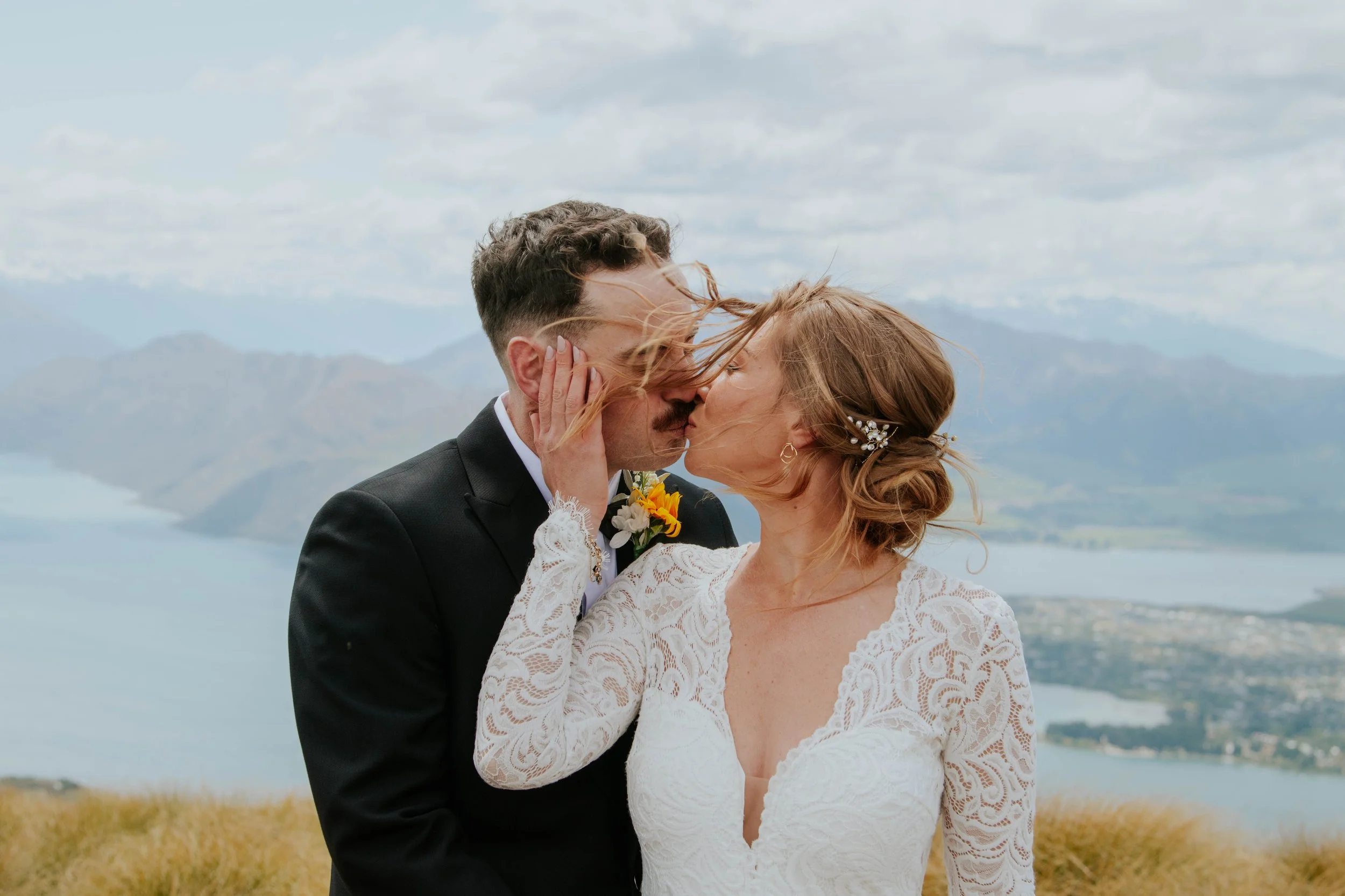 A couple in wedding attire sharing a kiss outdoors with mountains and a lake in the background.