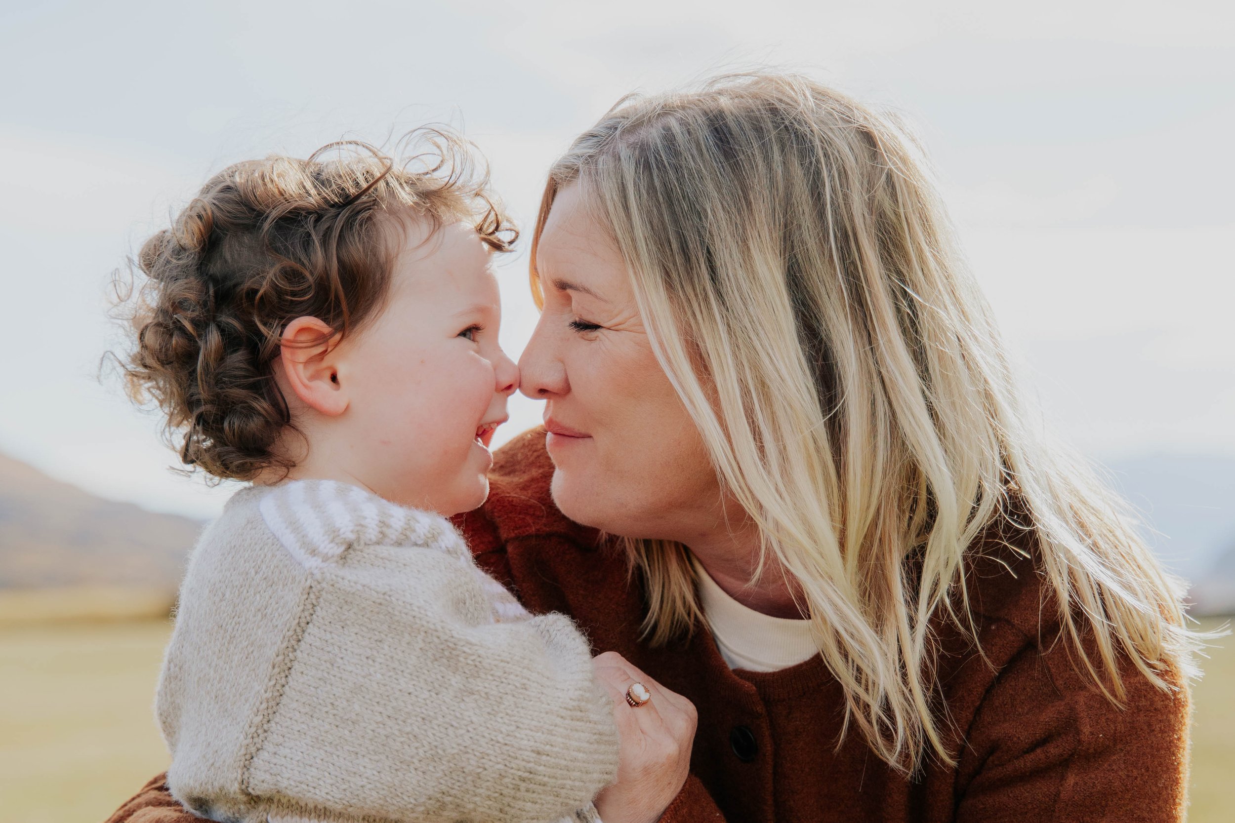 A woman and a young girl are touching noses, smiling and enjoying a moment outdoors, with a cloudy sky in the background.
