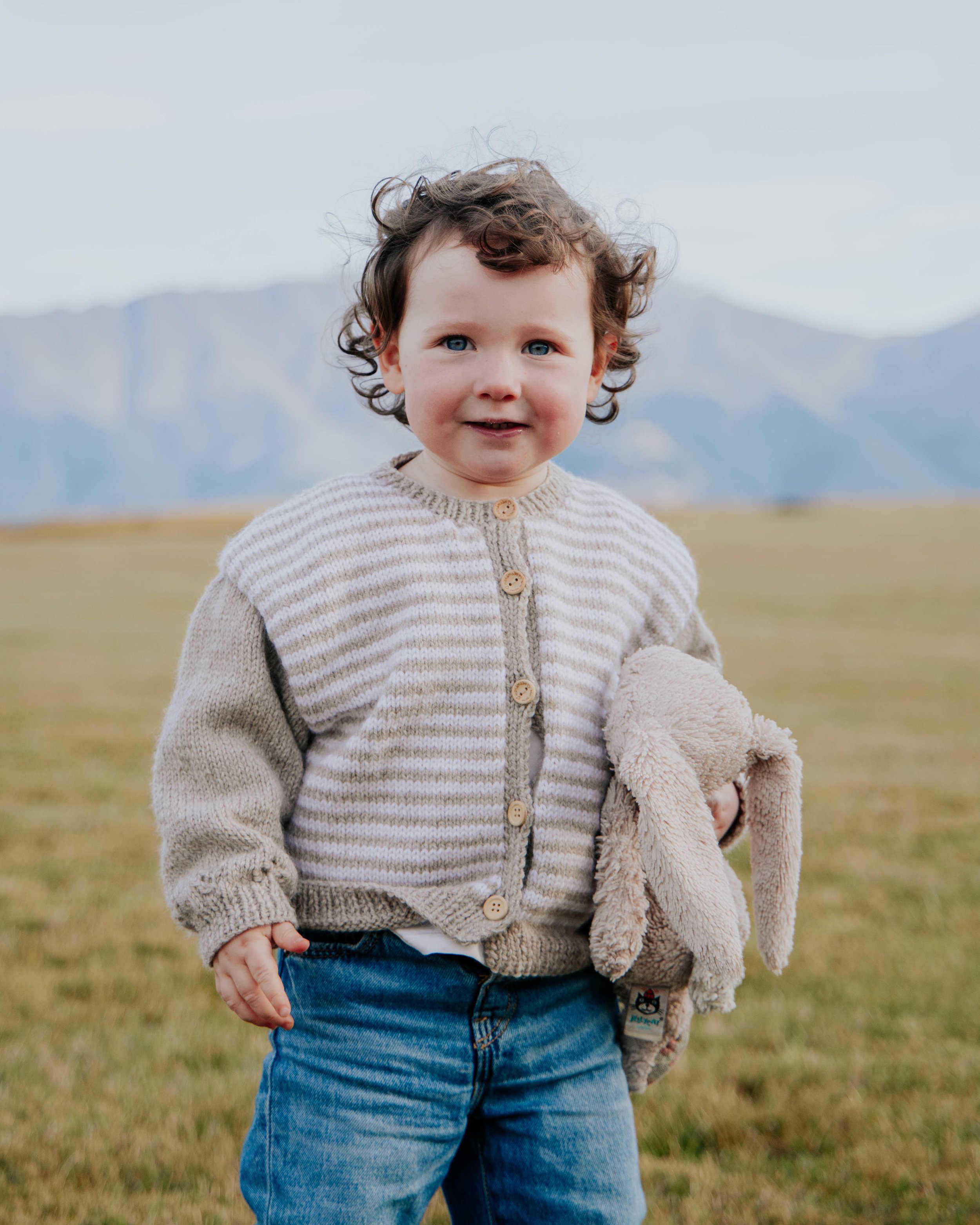 A young child with curly hair, blue eyes, and a slight smile, wearing a beige and brown striped sweater with buttons, standing outdoors on grass with mountainous terrain in the background and holding a plush teddy bear.