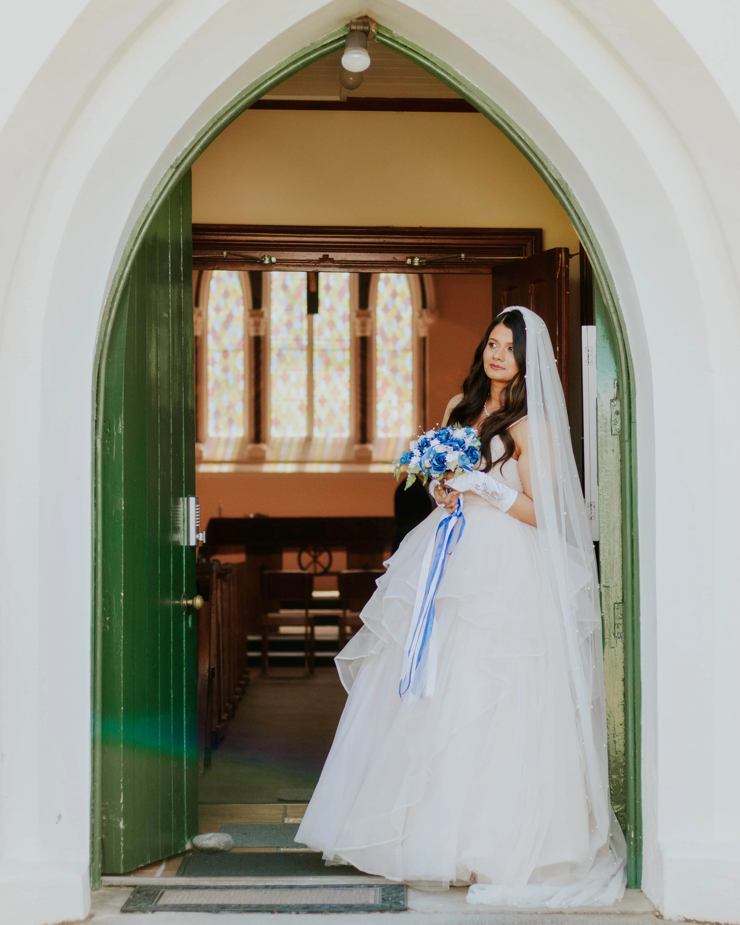 A bride in a white wedding gown holding a blue flower bouquet, standing in a doorway of a church with stained glass windows in the background.