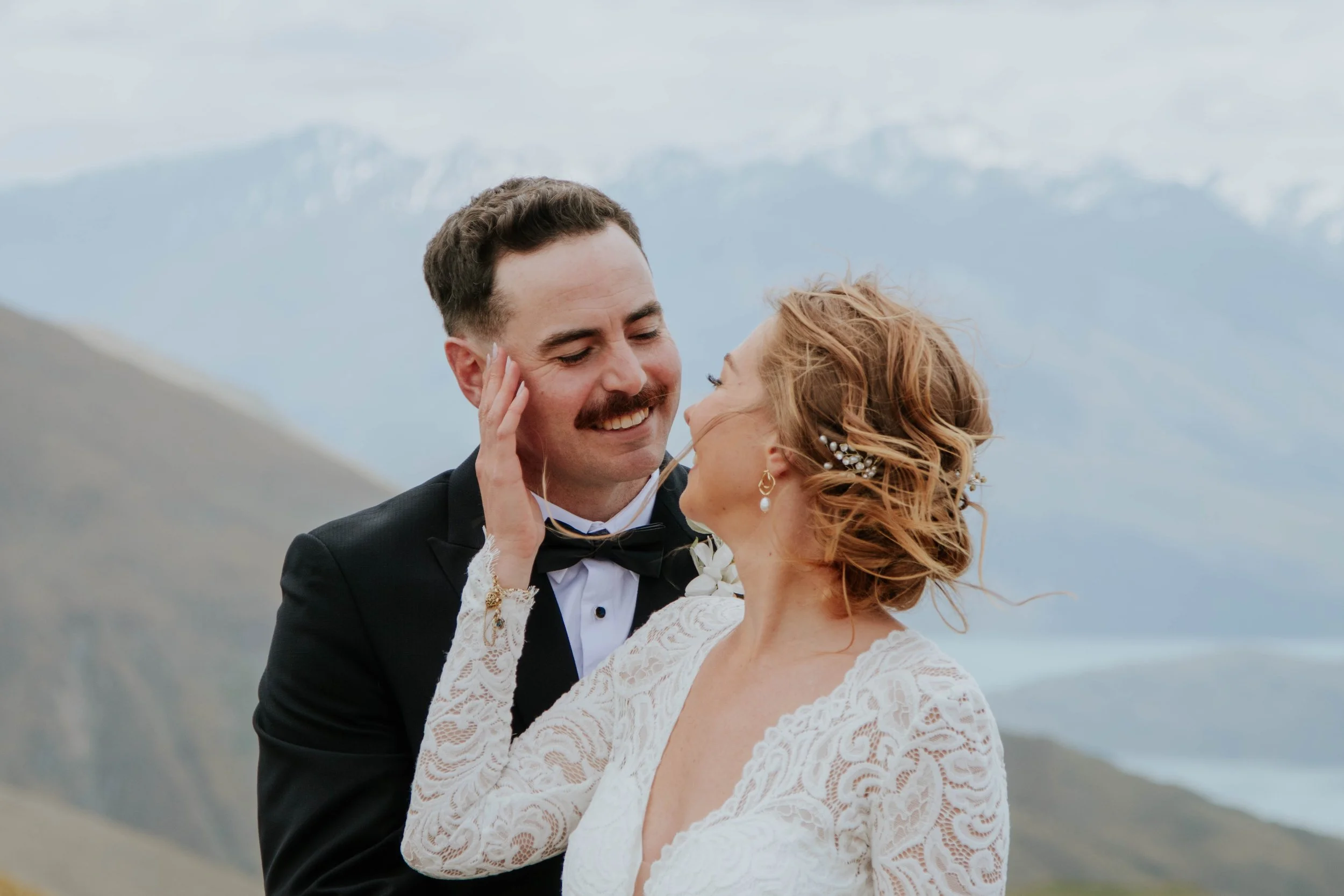 A newlywed couple standing outdoors with a mountain landscape in the background, sharing an intimate moment. The groom is wearing a black tuxedo with a bow tie, and the bride is in a lace wedding dress with her hair styled in loose curls adorned with