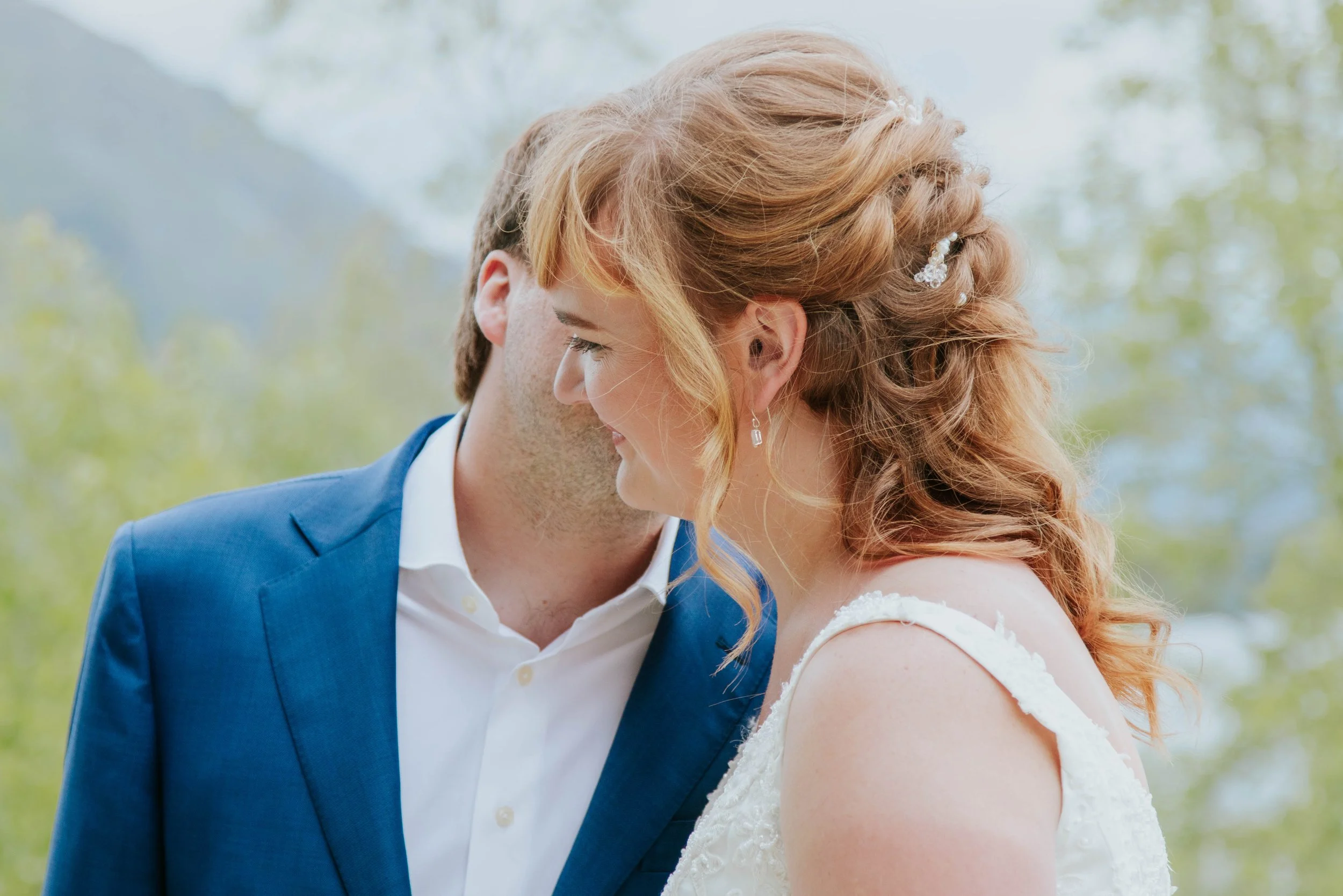 A bride and groom share a close moment, with foreheads touching and smiling, outdoors with a blurred natural background.