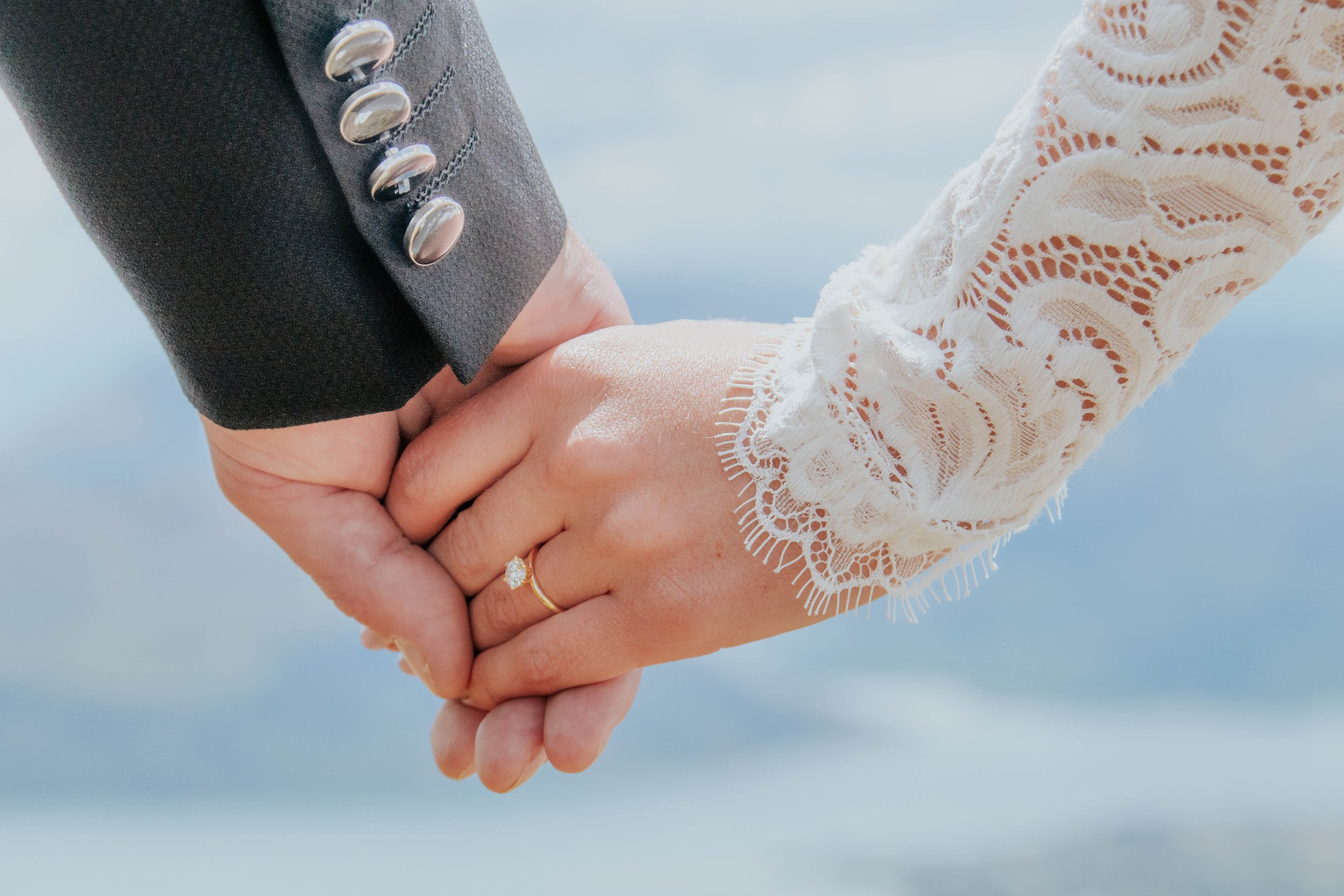 Close-up of a man and woman holding hands, with the woman wearing a lace long-sleeve shirt and engagement ring, and the man wearing a dark suit with silver buttons.