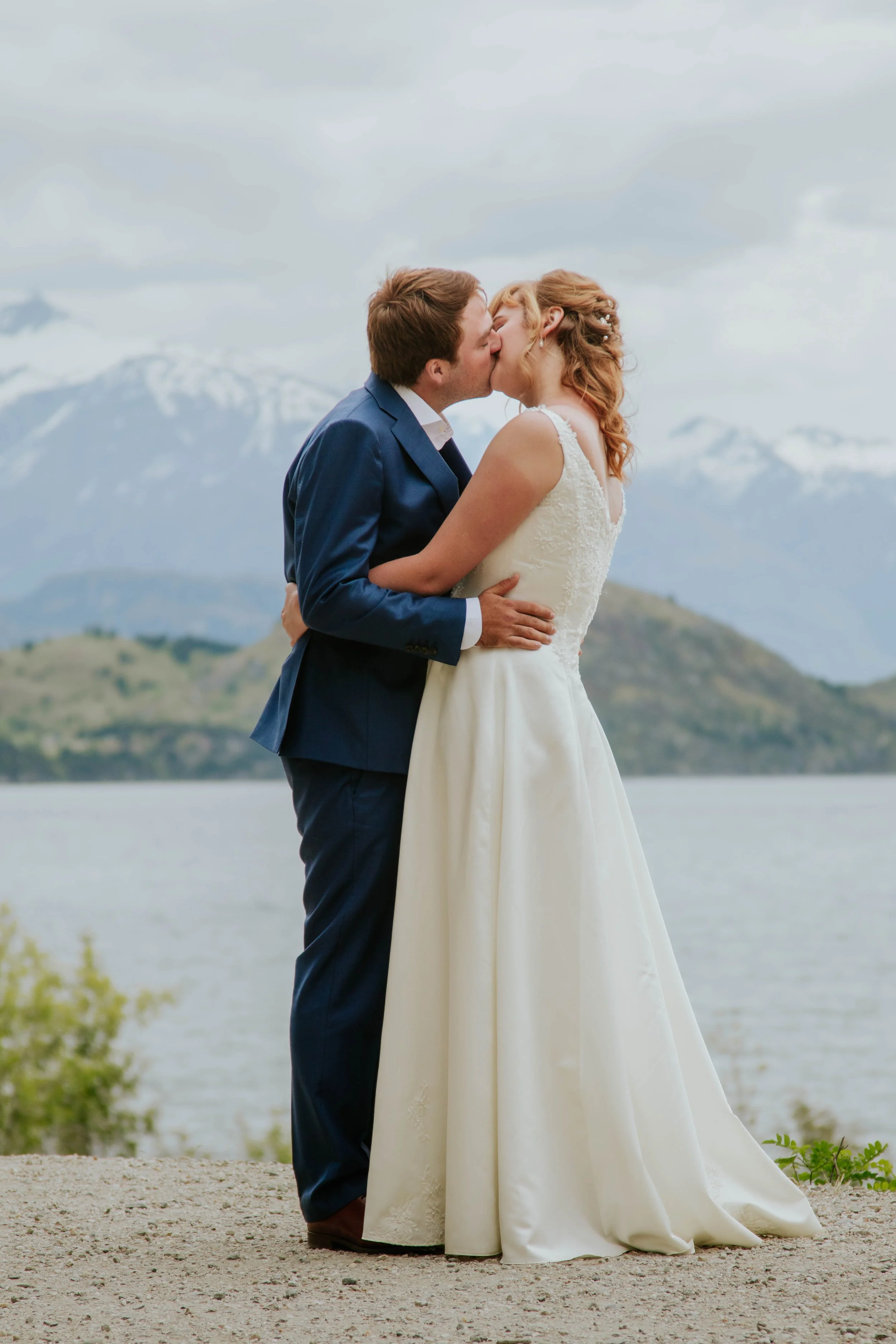 A bride and groom sharing a kiss outdoors near a lake with mountains in the background.