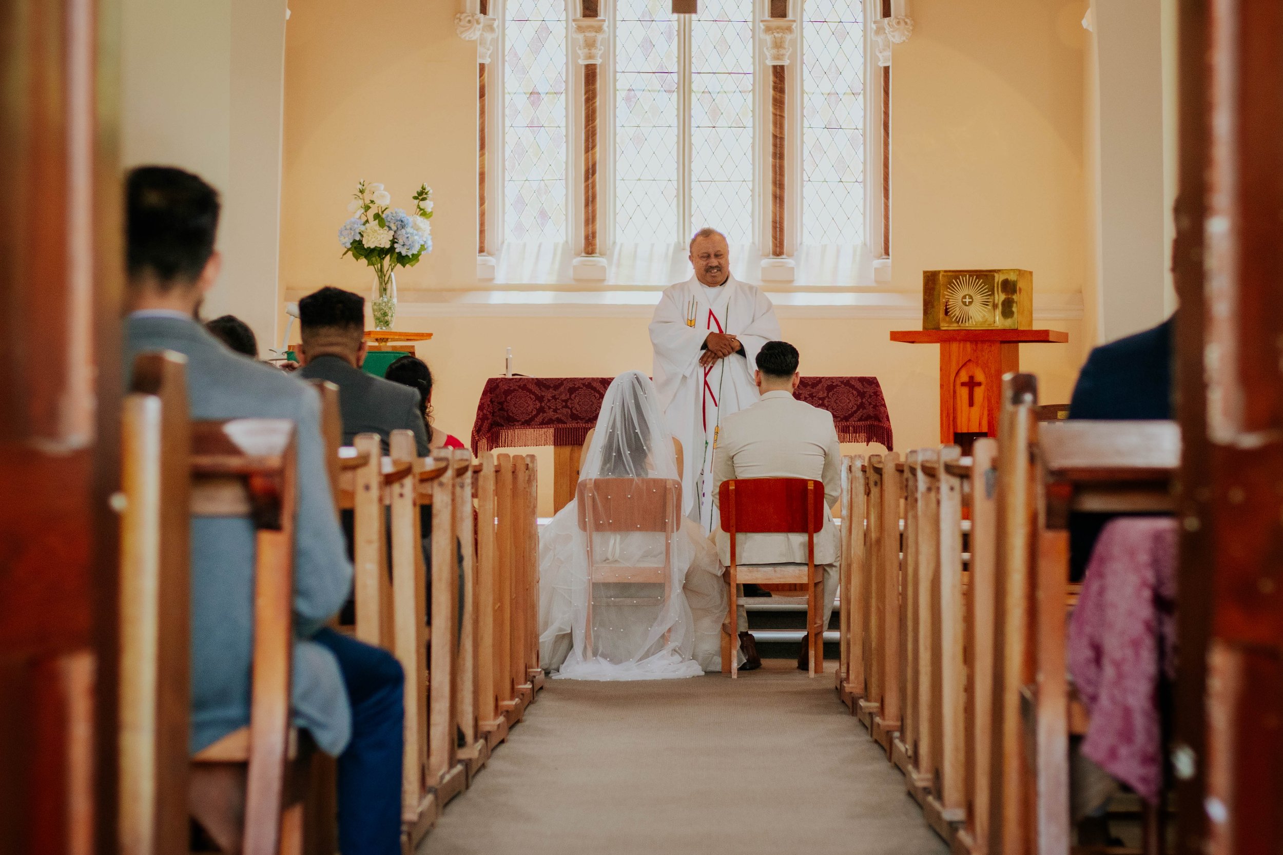 A wedding ceremony taking place in a church with the bride and groom sitting before a minister, who is speaking, and guests seated on wooden pews observing.