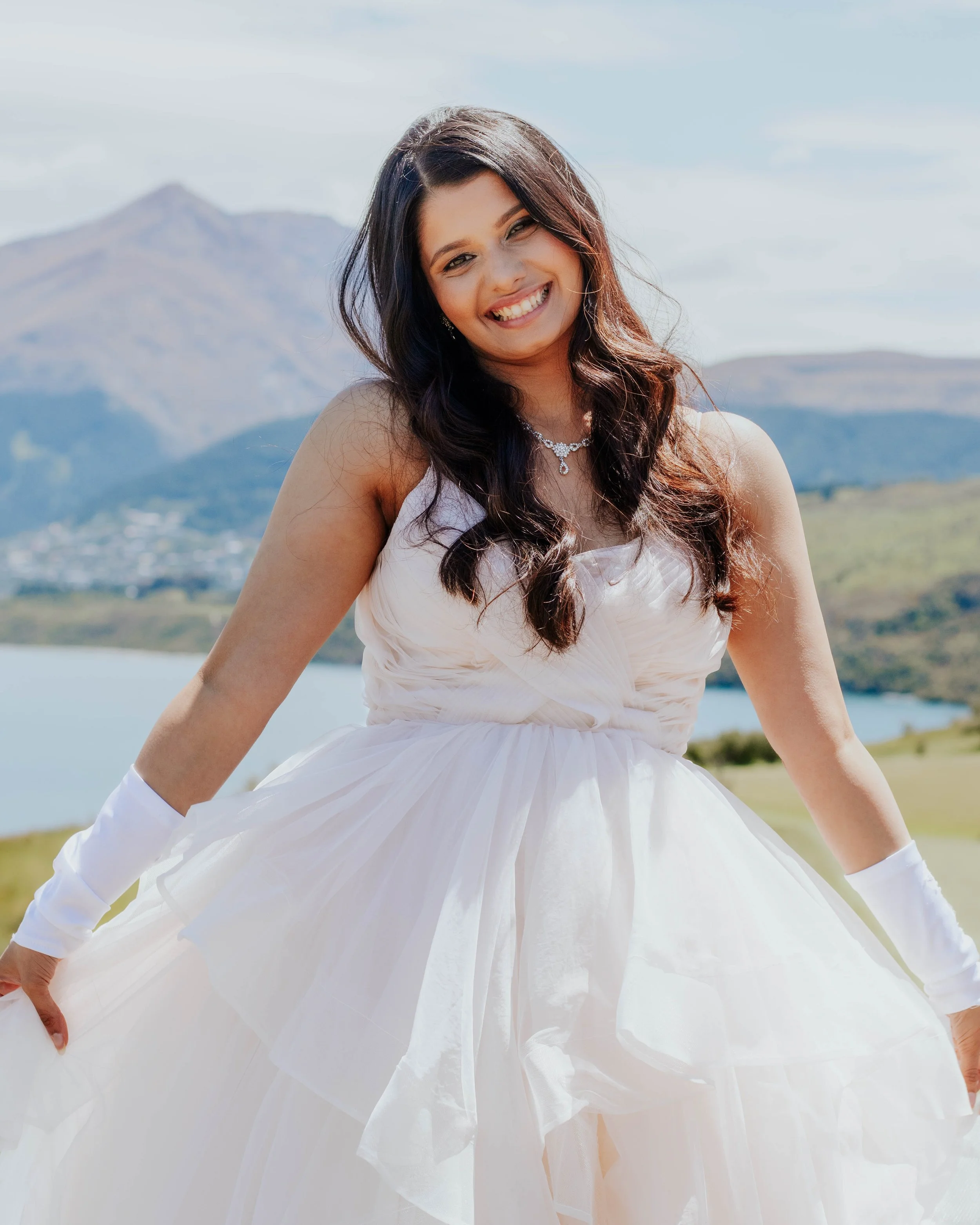 A woman in a white wedding dress standing outdoors with mountains and a lake in the background, smiling at the camera.