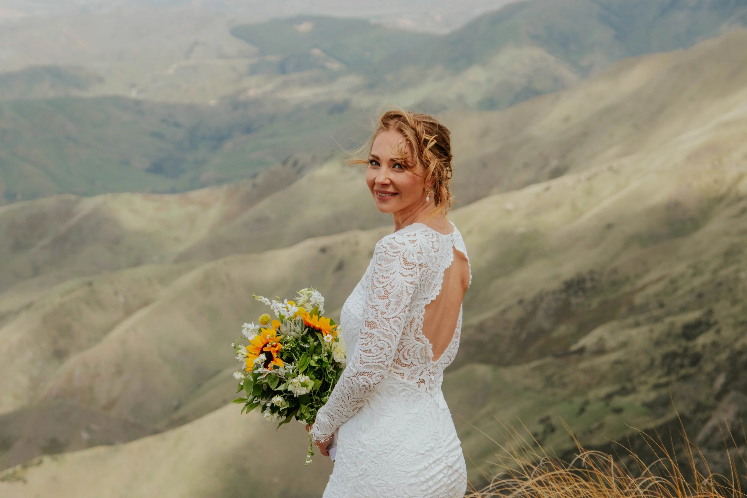 A woman in a white lace wedding dress holding a bouquet of yellow, white, and green flowers, standing outdoors with rolling green hills in the background.